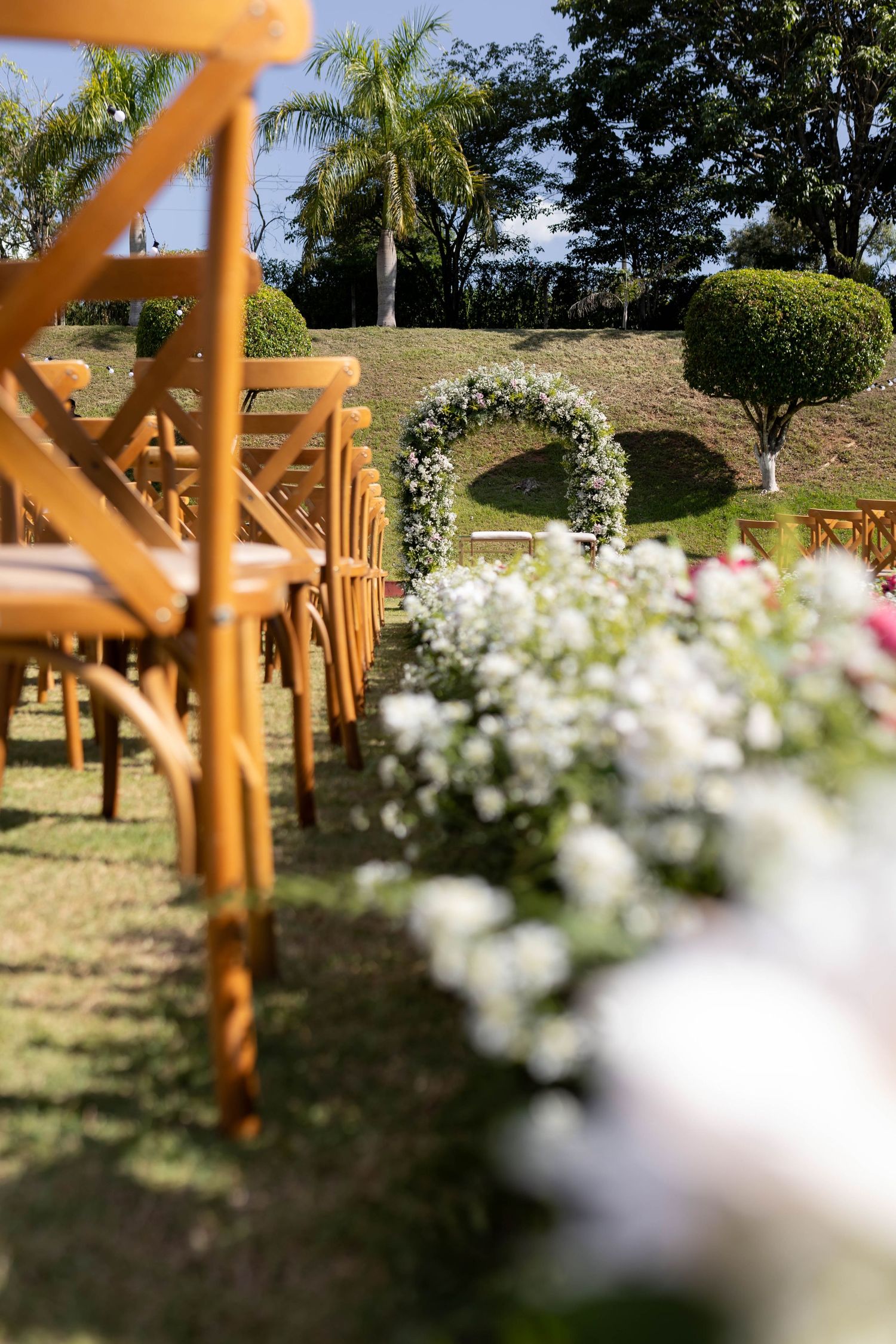 Outdoor wedding ceremony setup with wooden cross-back chairs, white floral aisle decorations, and a circular flower arch on green lawn.