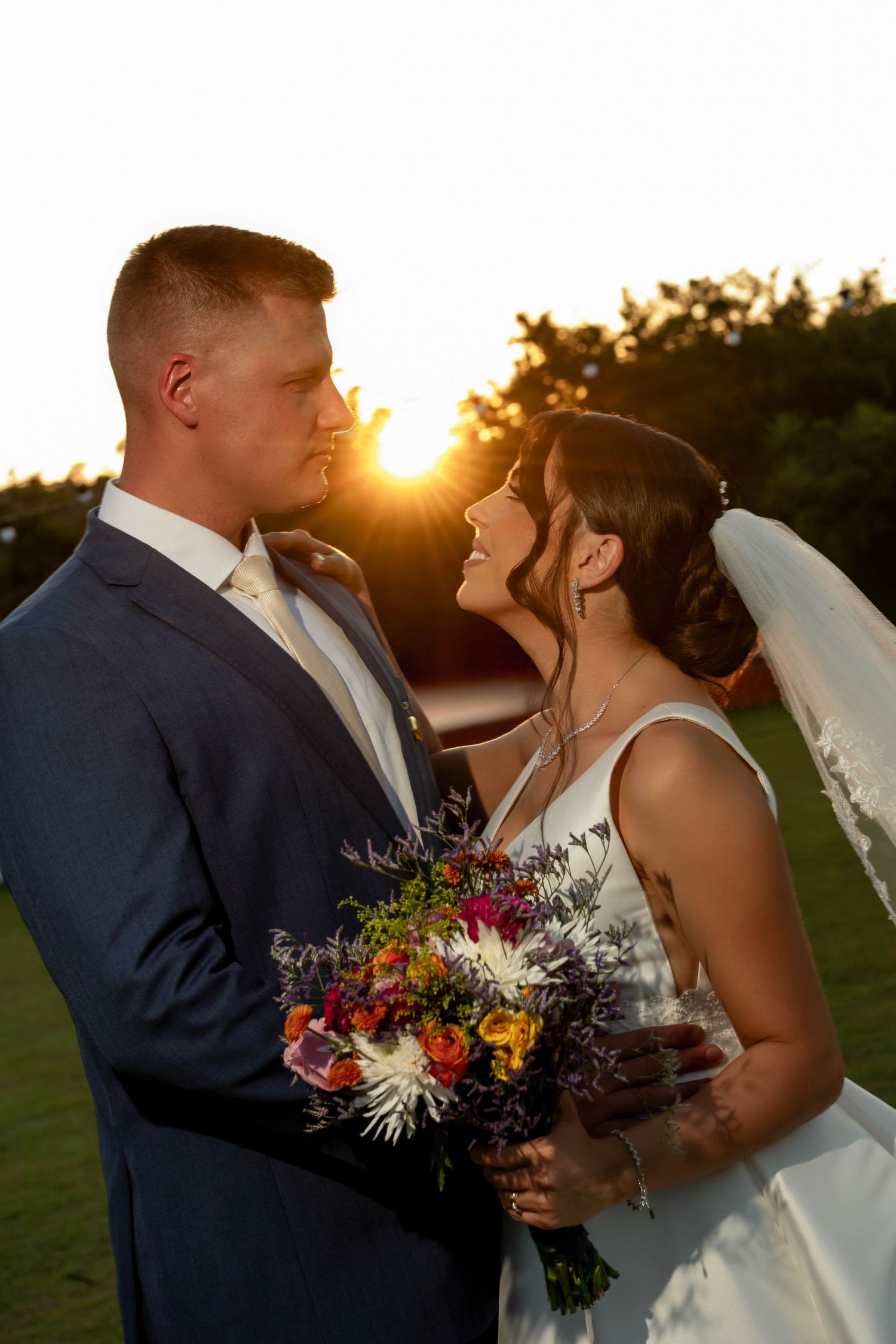 Bride and groom share a romantic sunset moment, she holds a colorful bouquet, golden light glowing behind them.