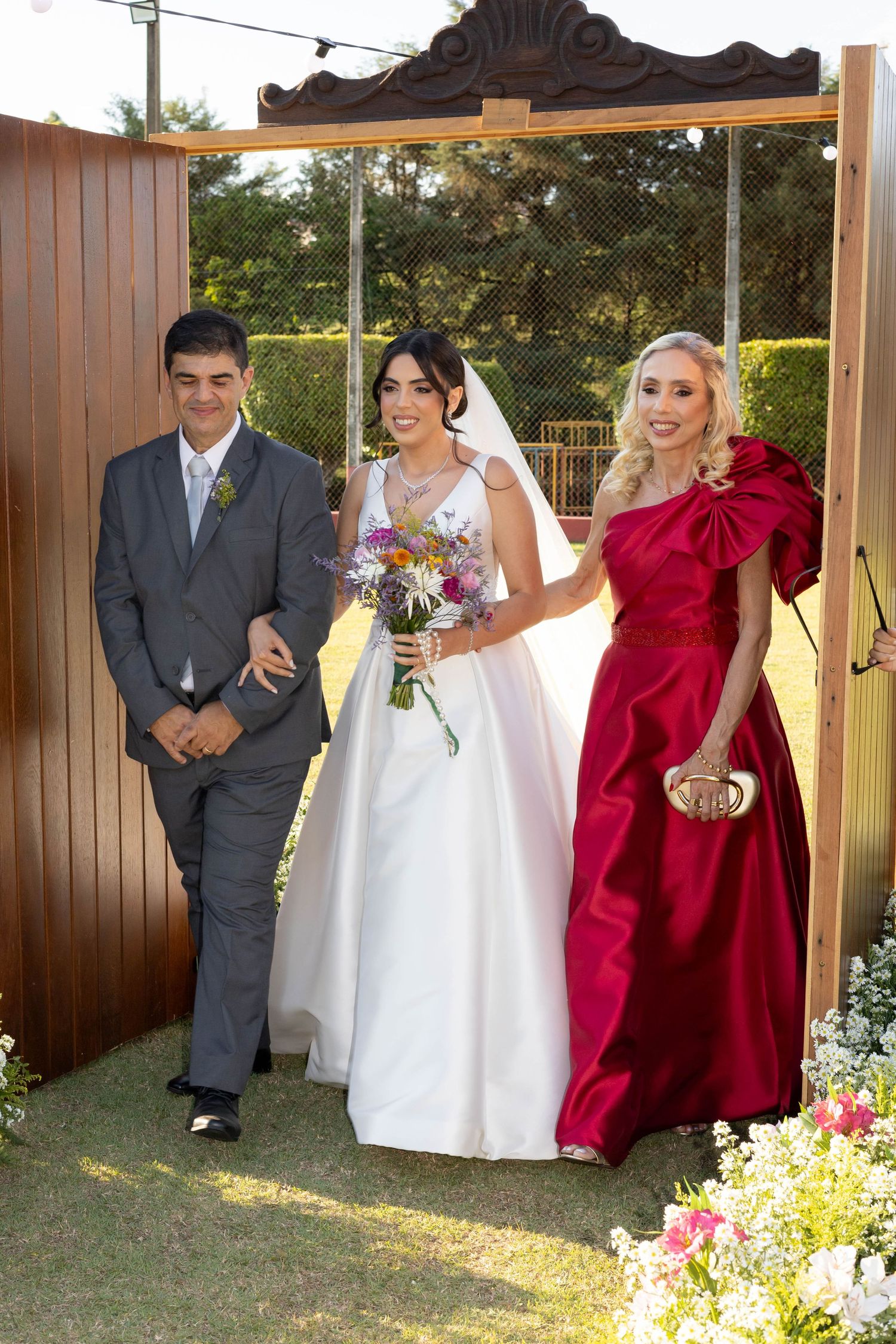 Bride in white gown with bouquet walks with man in gray suit and woman in red dress at outdoor wedding ceremony.