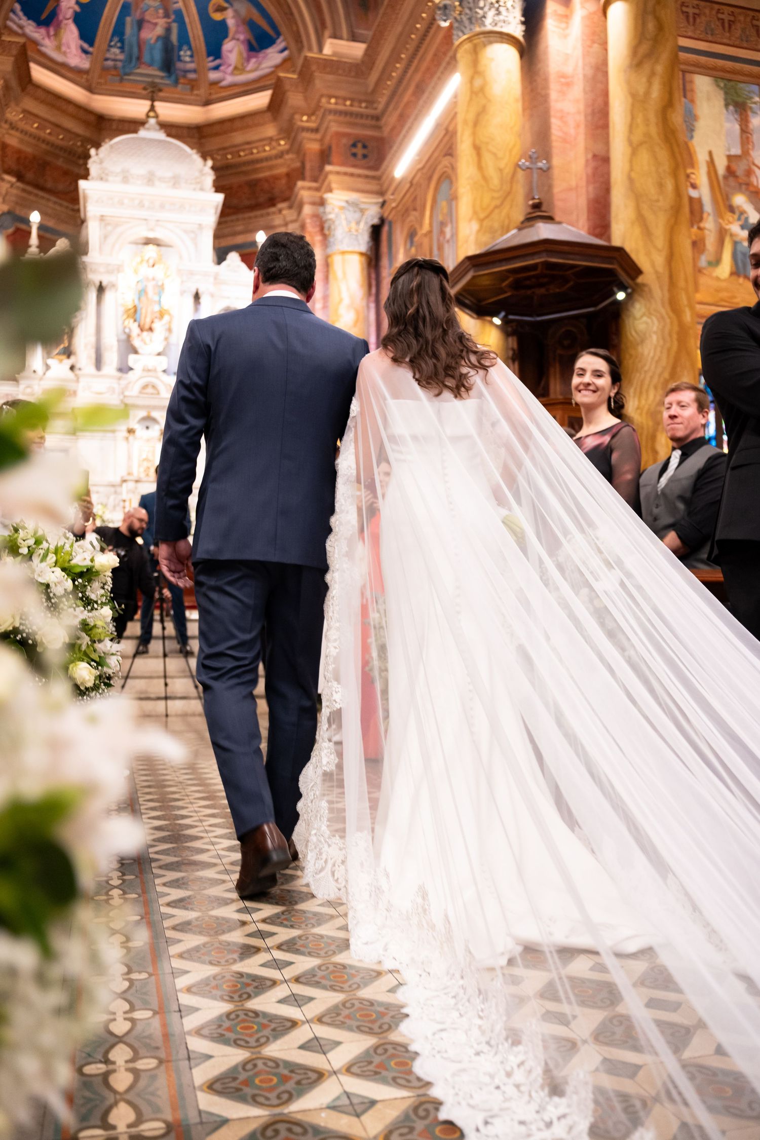 Bride in white gown with long veil walks down church aisle with groom in navy suit during wedding ceremony.