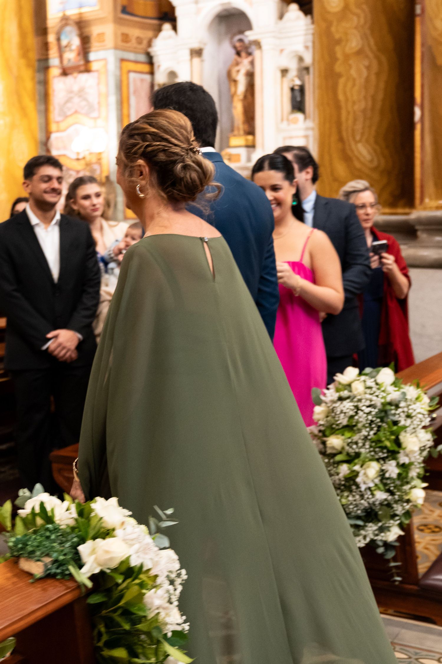 Elegant wedding ceremony in a church with guests in formal attire, including a woman in a sage green cape dress.