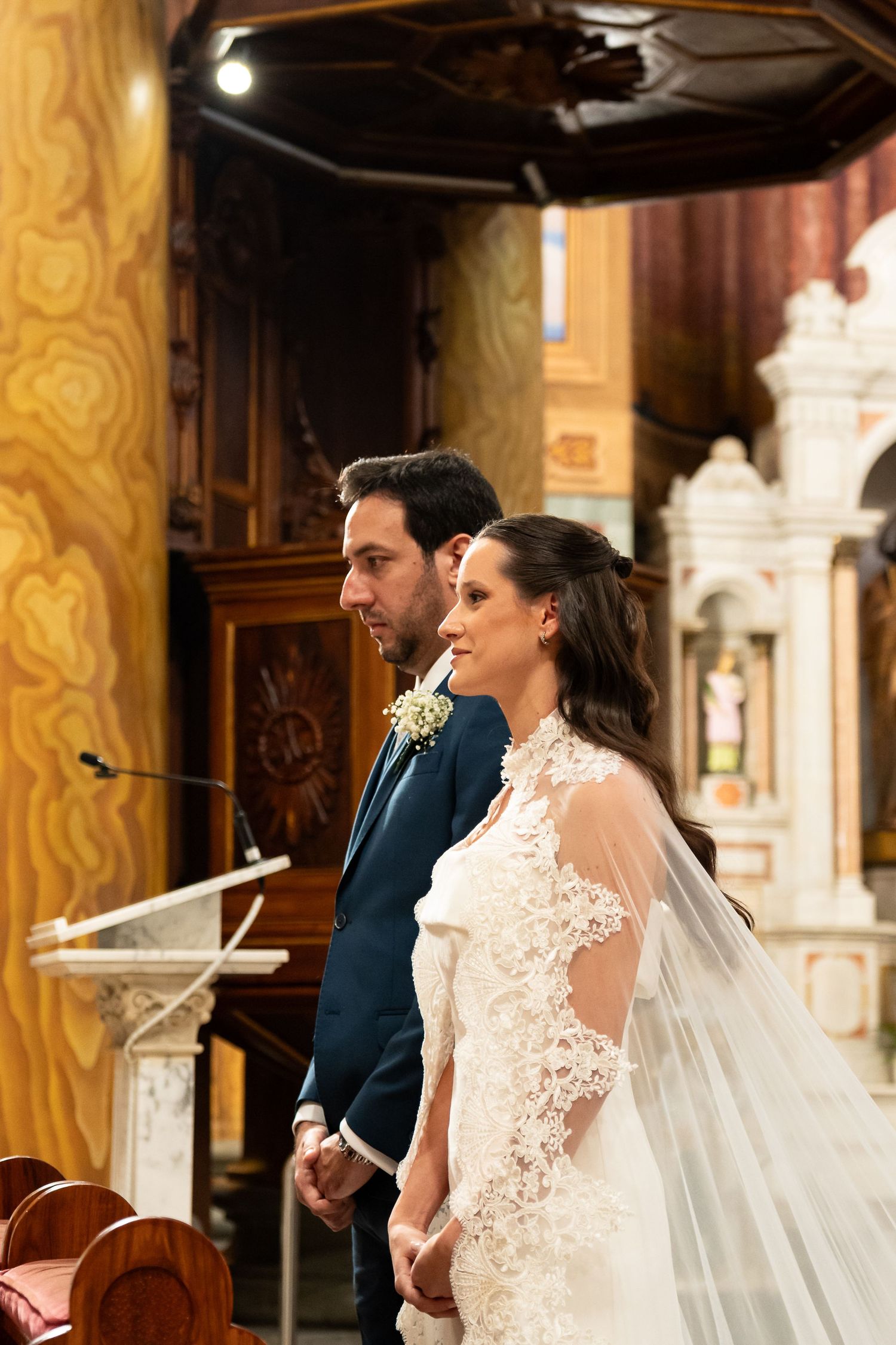Bride and groom stand together at church altar during wedding ceremony, bride wearing lace gown with veil.
