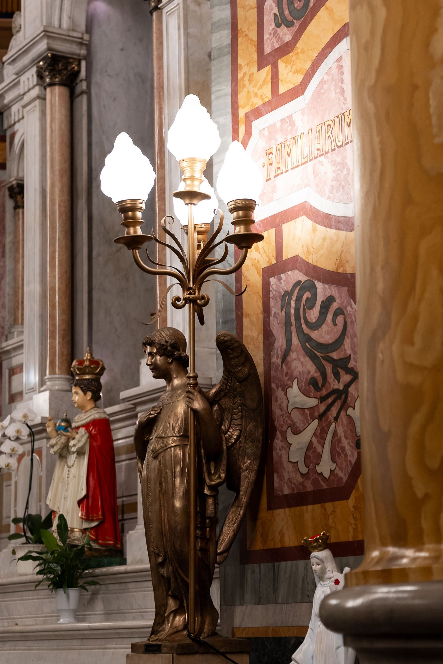 Bronze angel candelabra with lit lamps inside an ornate church with marble walls and religious statues.