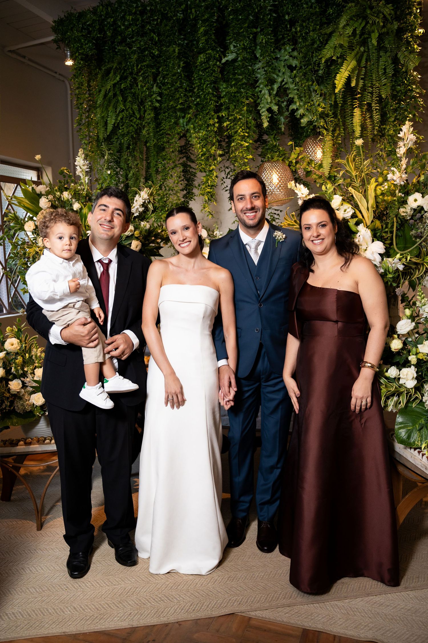 Wedding party posing in front of lush green floral backdrop, bride in white gown with groom and guests.