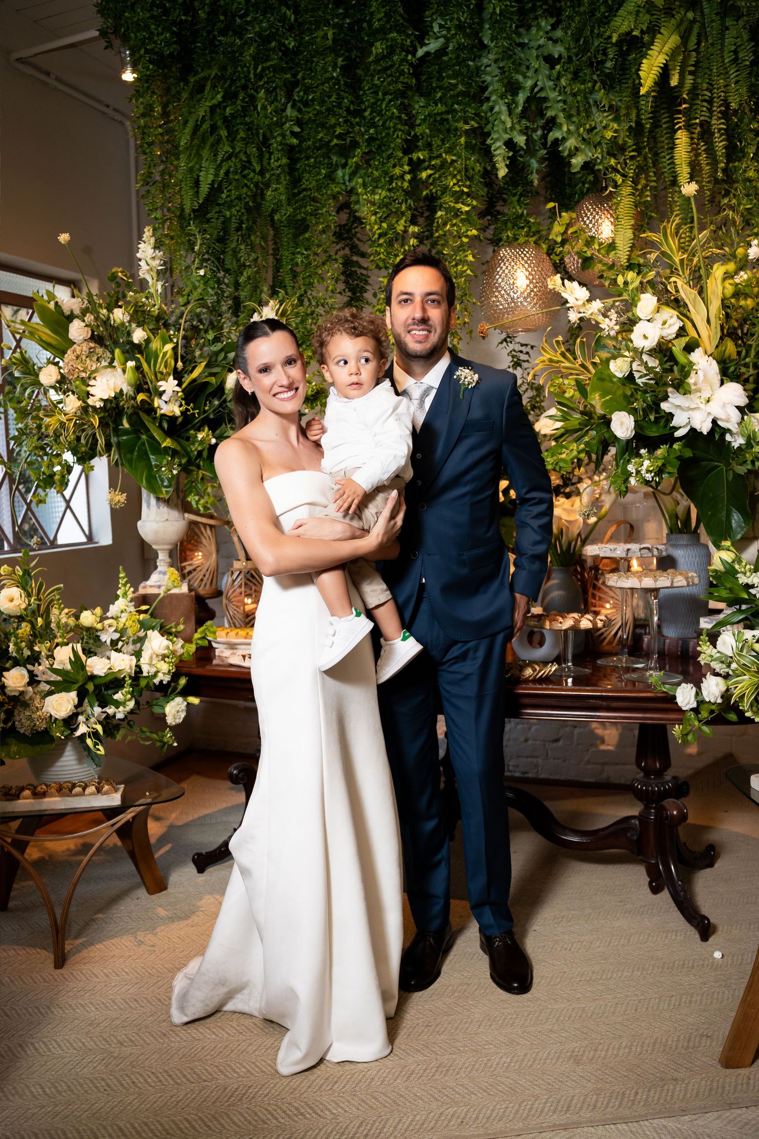 Bride in white gown and groom in navy suit pose with toddler at elegant wedding reception with floral greenery backdrop.