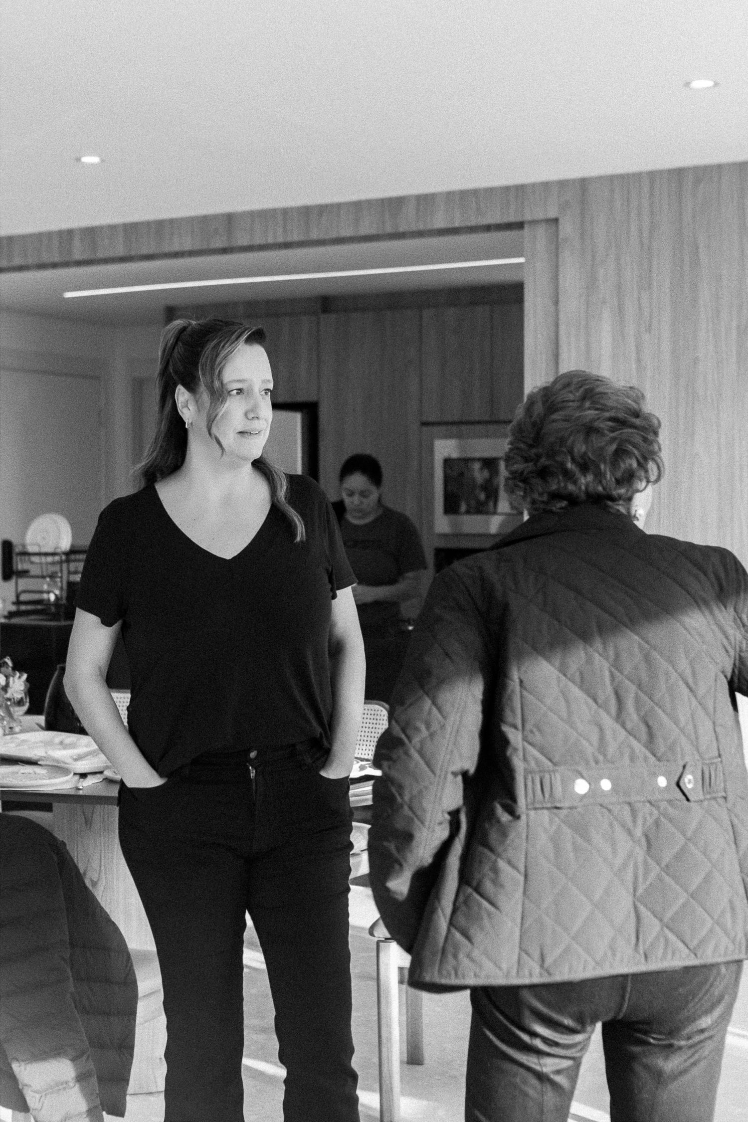 Black and white photo of two people conversing indoors, woman in black t-shirt facing man in quilted jacket.