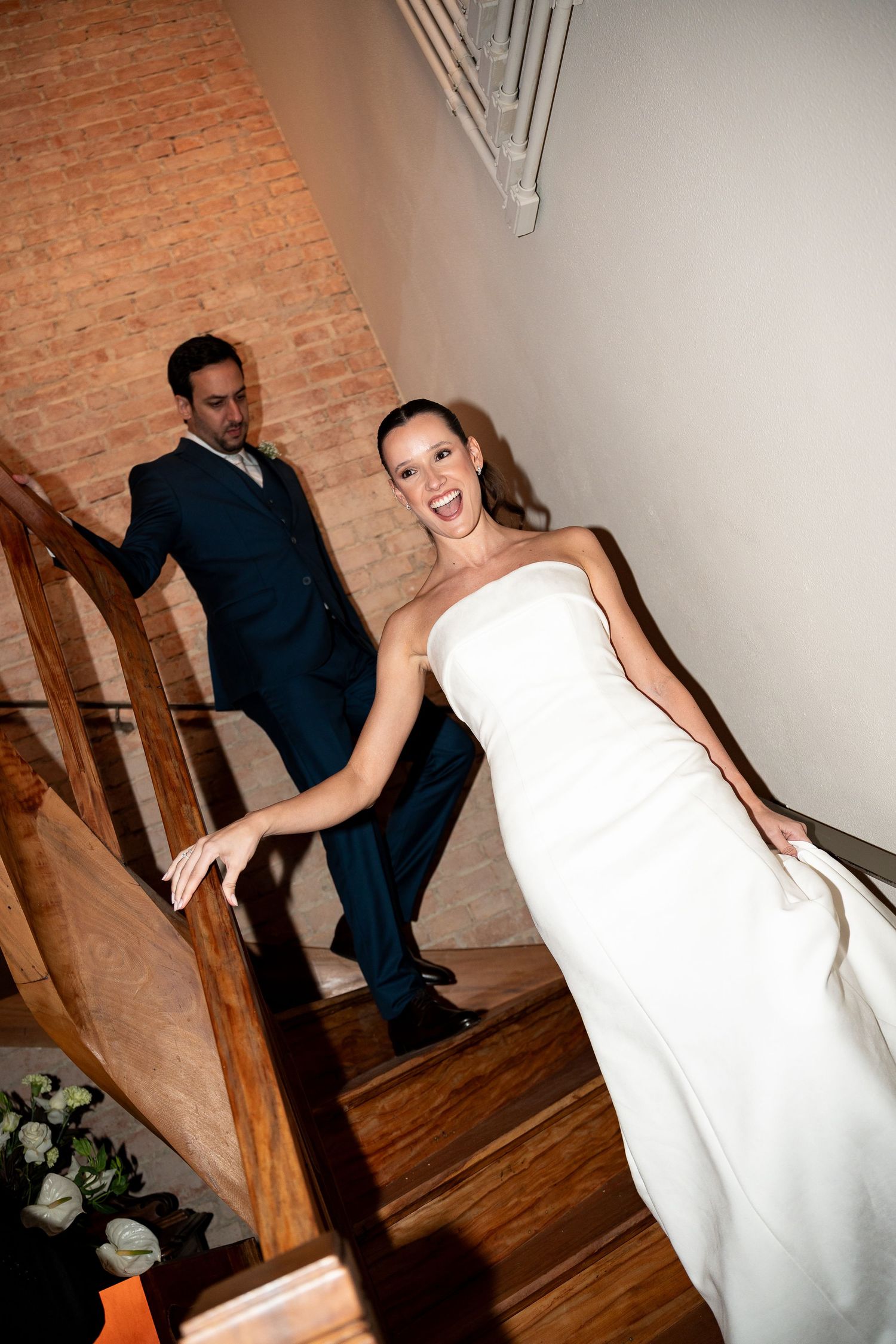 Bride in white strapless gown laughing on wooden staircase with groom in navy suit behind her.