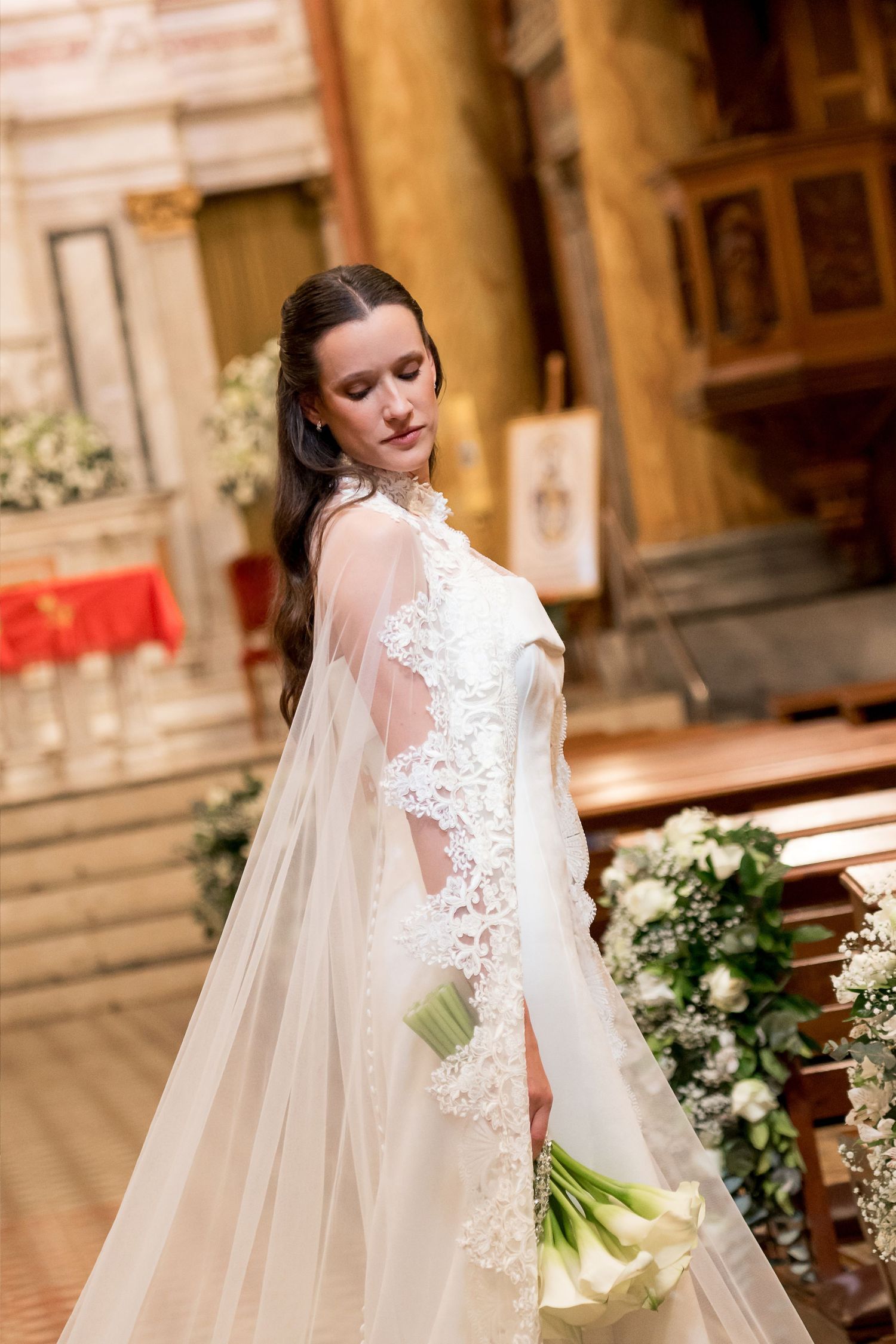Elegant bride in lace wedding gown holding white bouquet inside ornate church with floral decorations.