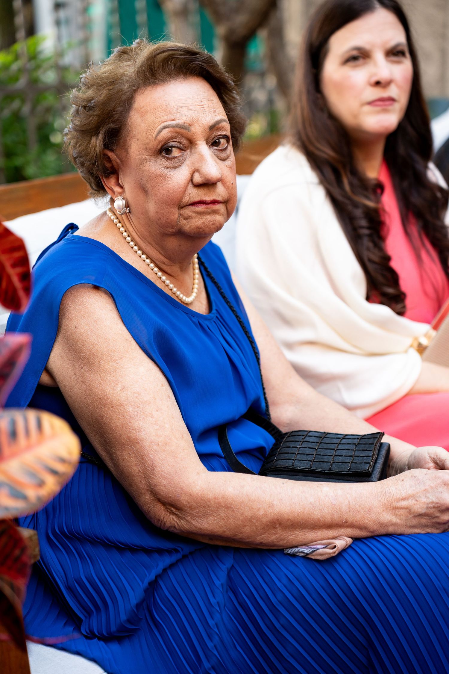 Elderly woman in blue dress with pearl necklace holding black clutch seated outdoors at formal event.