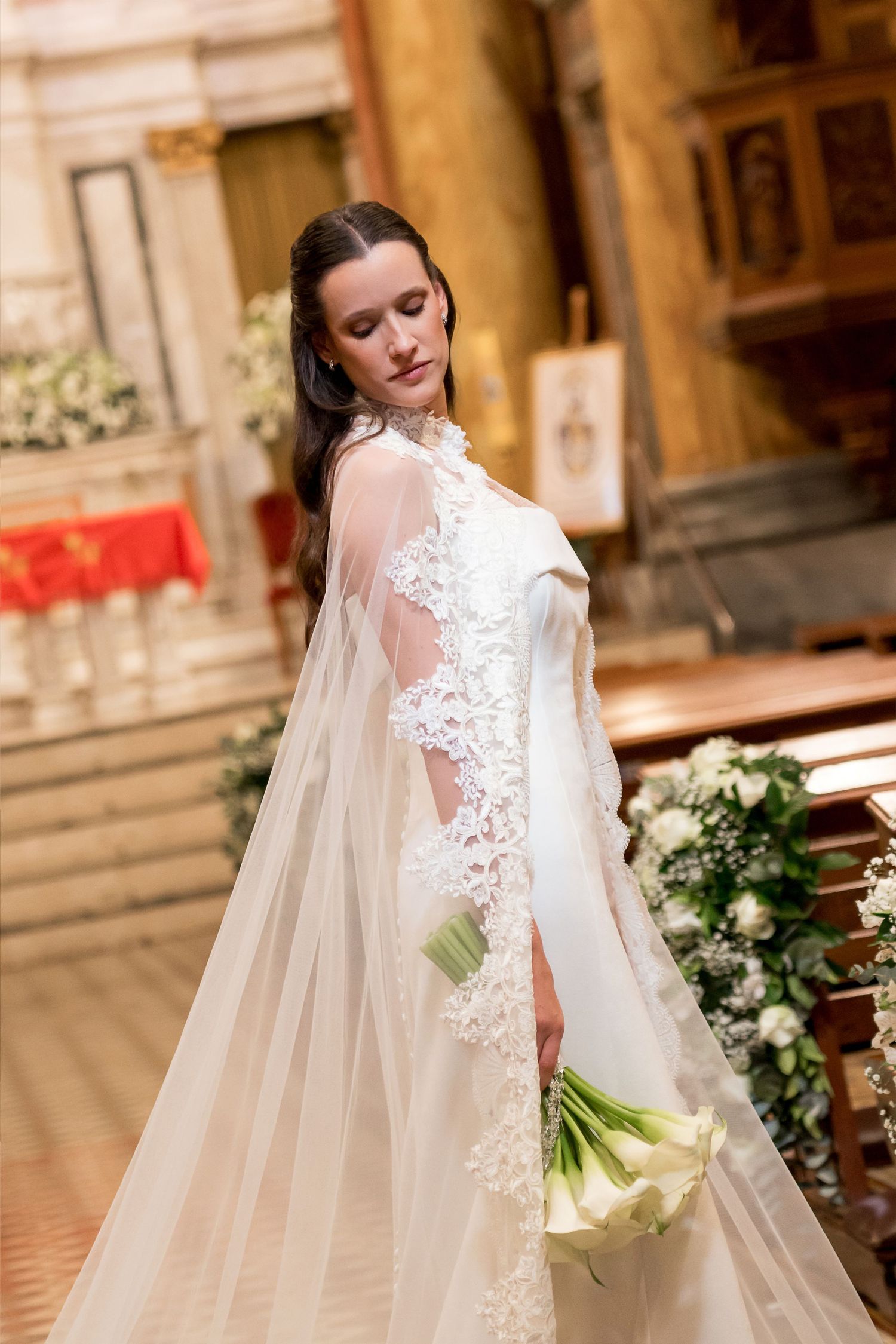 Bride in elegant lace wedding gown with sheer cape holds white calla lily bouquet inside ornate church.