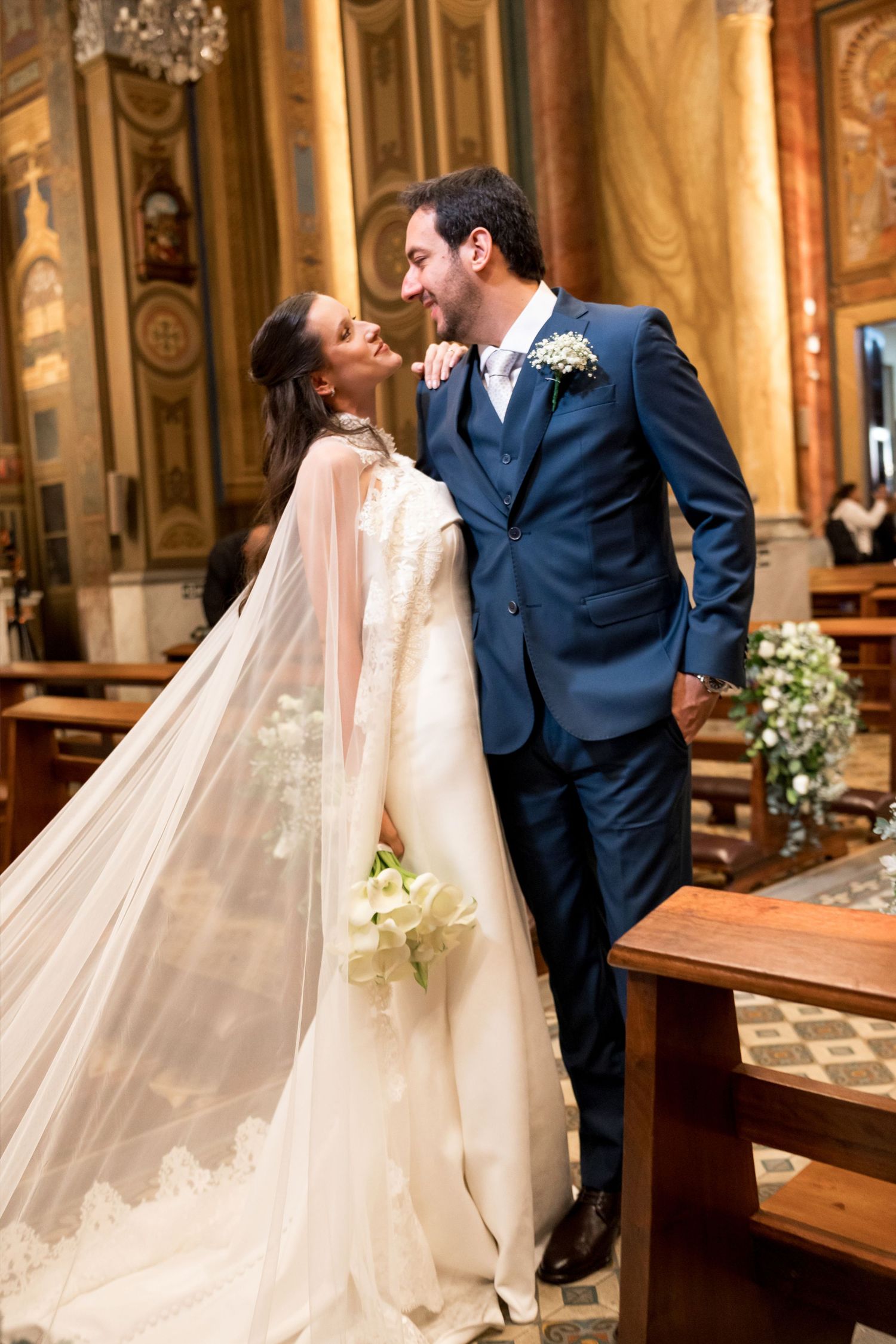 Bride in white gown with veil and groom in navy suit share a romantic moment in an elegant church.