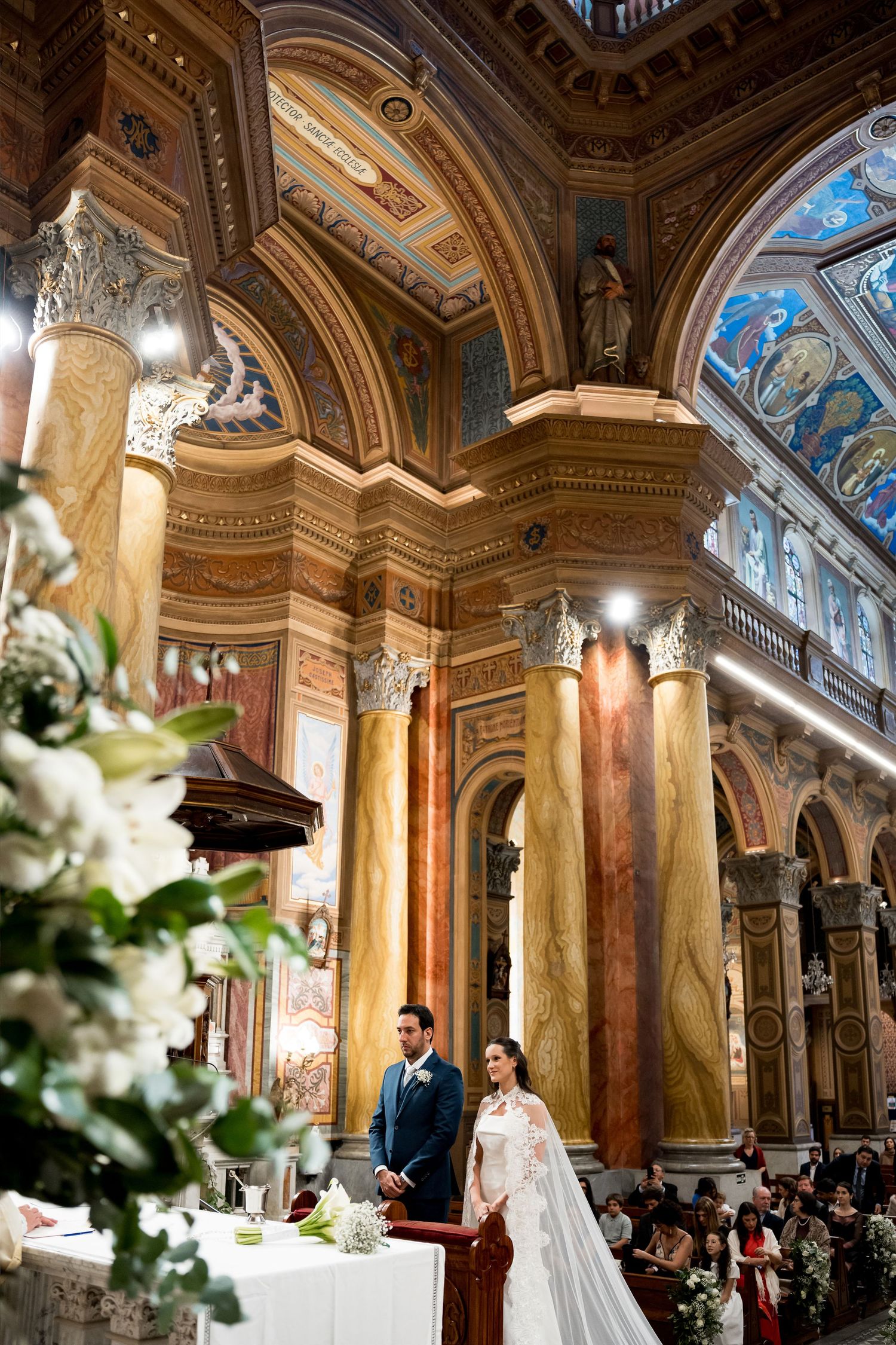 Bride and groom stand at the altar of an ornate church with gilded arches, marble columns, and colorful frescoes.