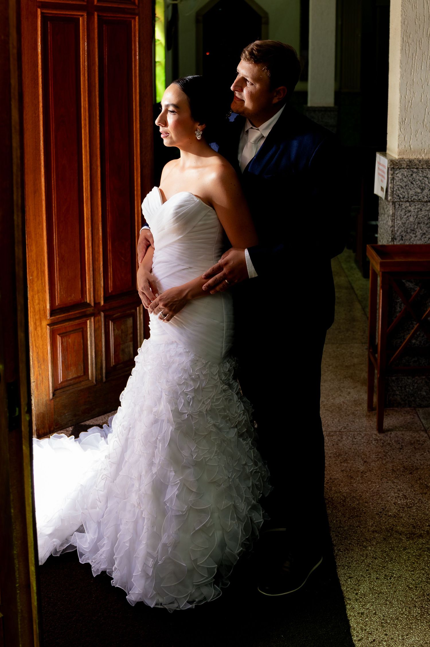 Bride in strapless mermaid gown embraced by groom in navy suit, standing by wooden doors in elegant venue.