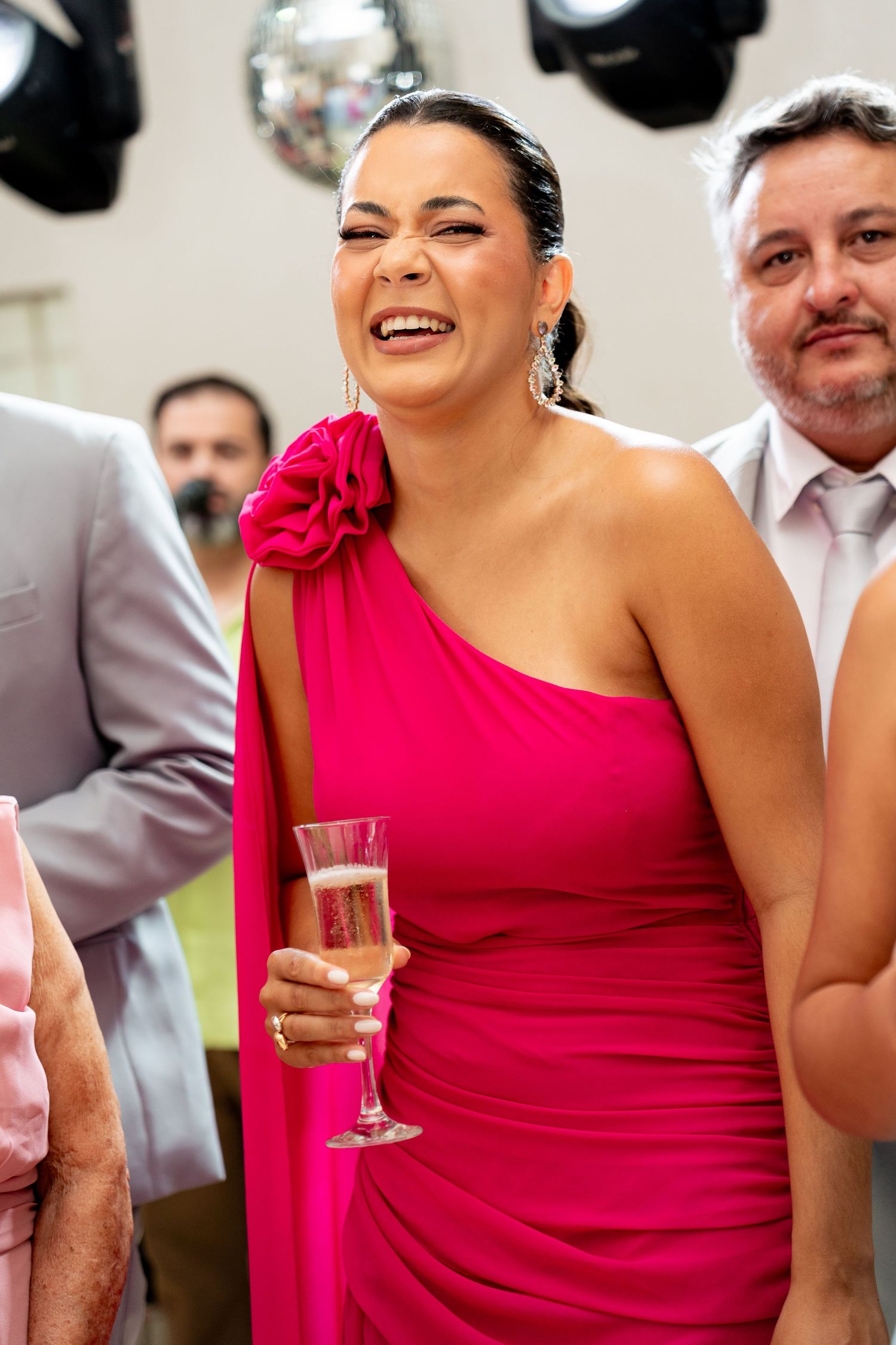 Woman in hot pink one-shoulder gown laughing joyfully, holding champagne flute at elegant celebration event.