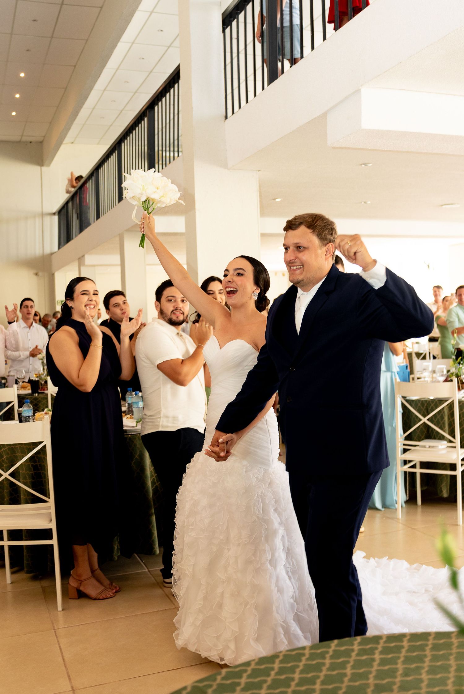 Joyful newlyweds celebrate their wedding reception entrance, bride in ruffled gown raising bouquet, groom in navy suit.