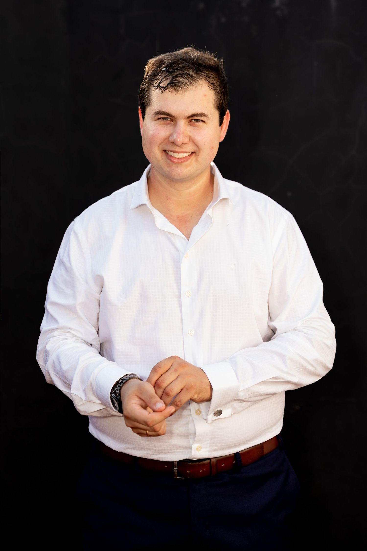 Smiling man in white dress shirt and brown belt posing against dark background.
