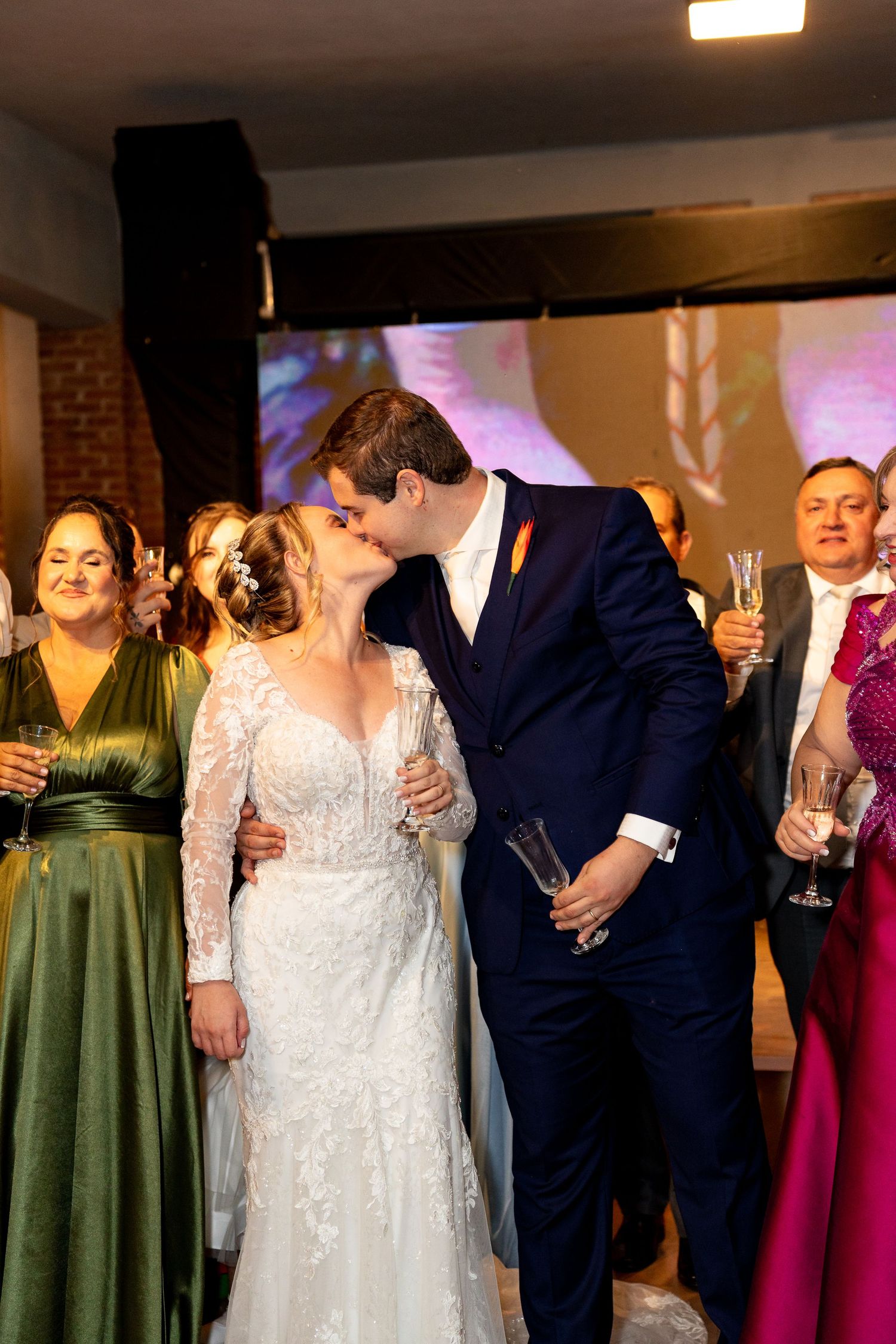 Bride and groom share a kiss at their wedding reception surrounded by guests in formal attire.