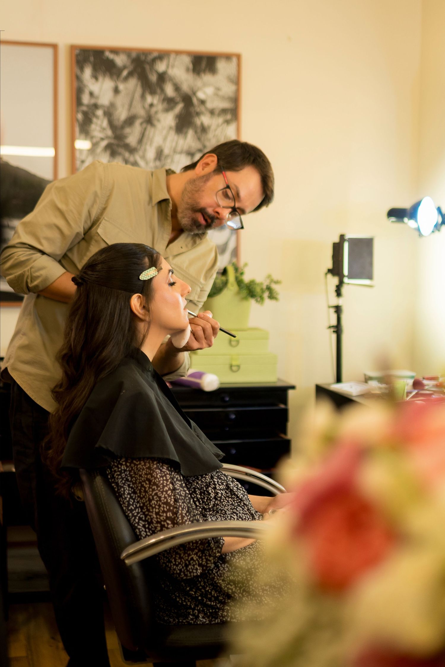 Male makeup artist applying lipstick to a woman seated in a salon chair in a warmly lit beauty studio.