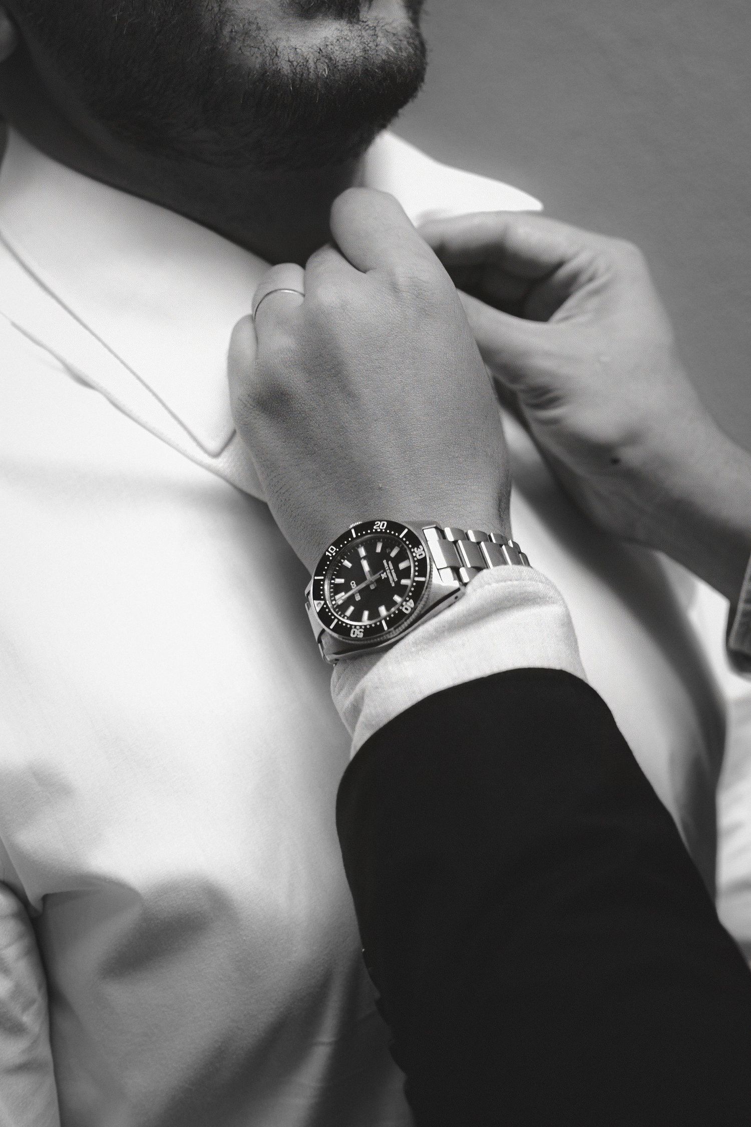 Black and white photo of a man in a suit adjusting his collar, wearing a luxury silver watch on his wrist.