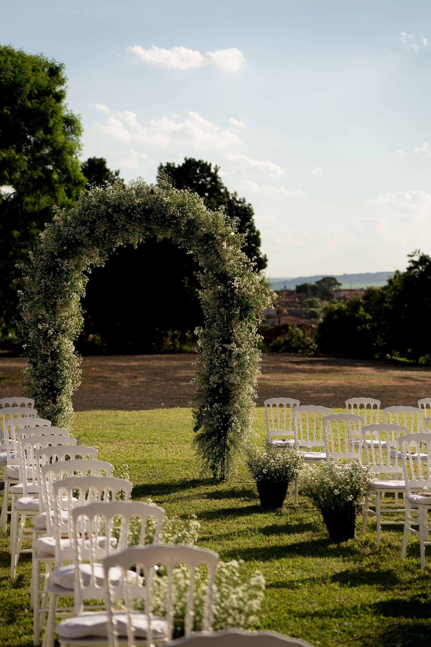 Outdoor wedding ceremony setup with white chairs, floral arch, and lush greenery under a blue sky.