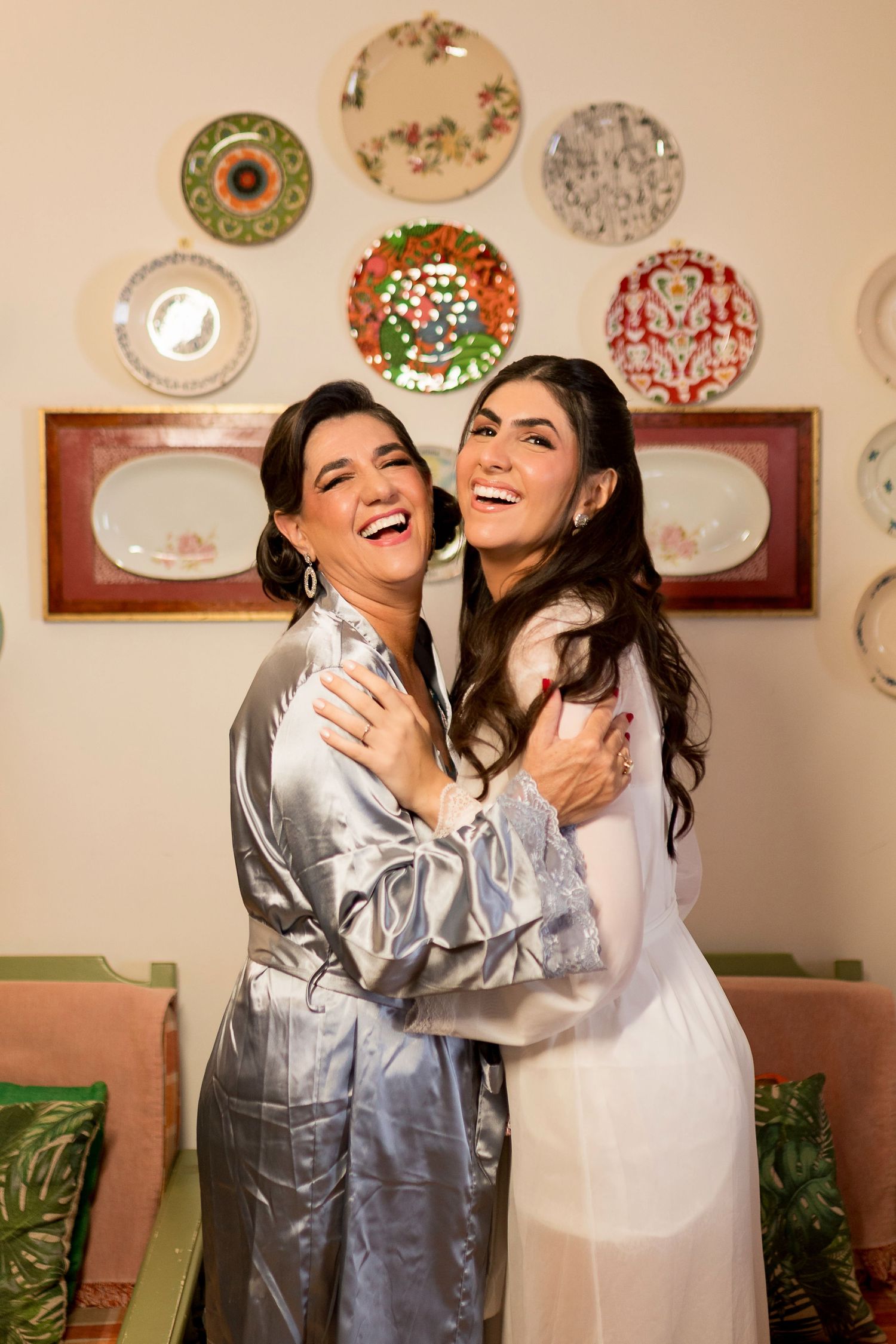 Two women in satin robes laughing and embracing joyfully in front of decorative wall plates.