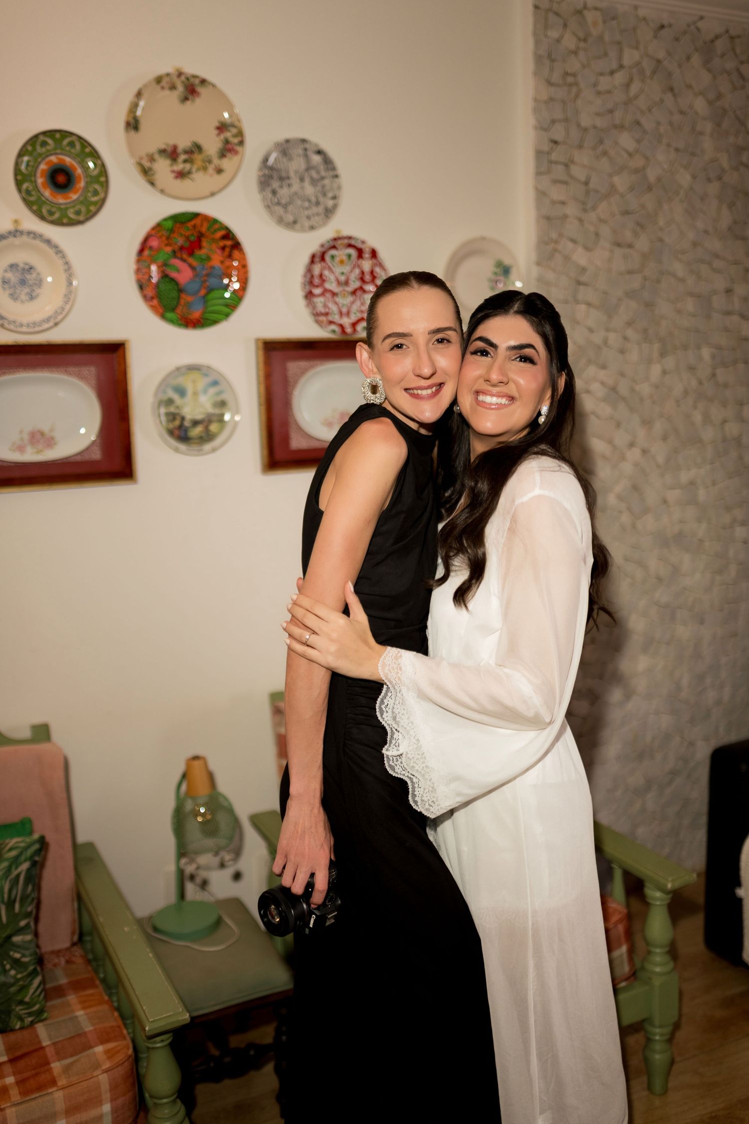 Two women hugging and smiling, one in a black dress and one in a white gown, standing in a decorated room.