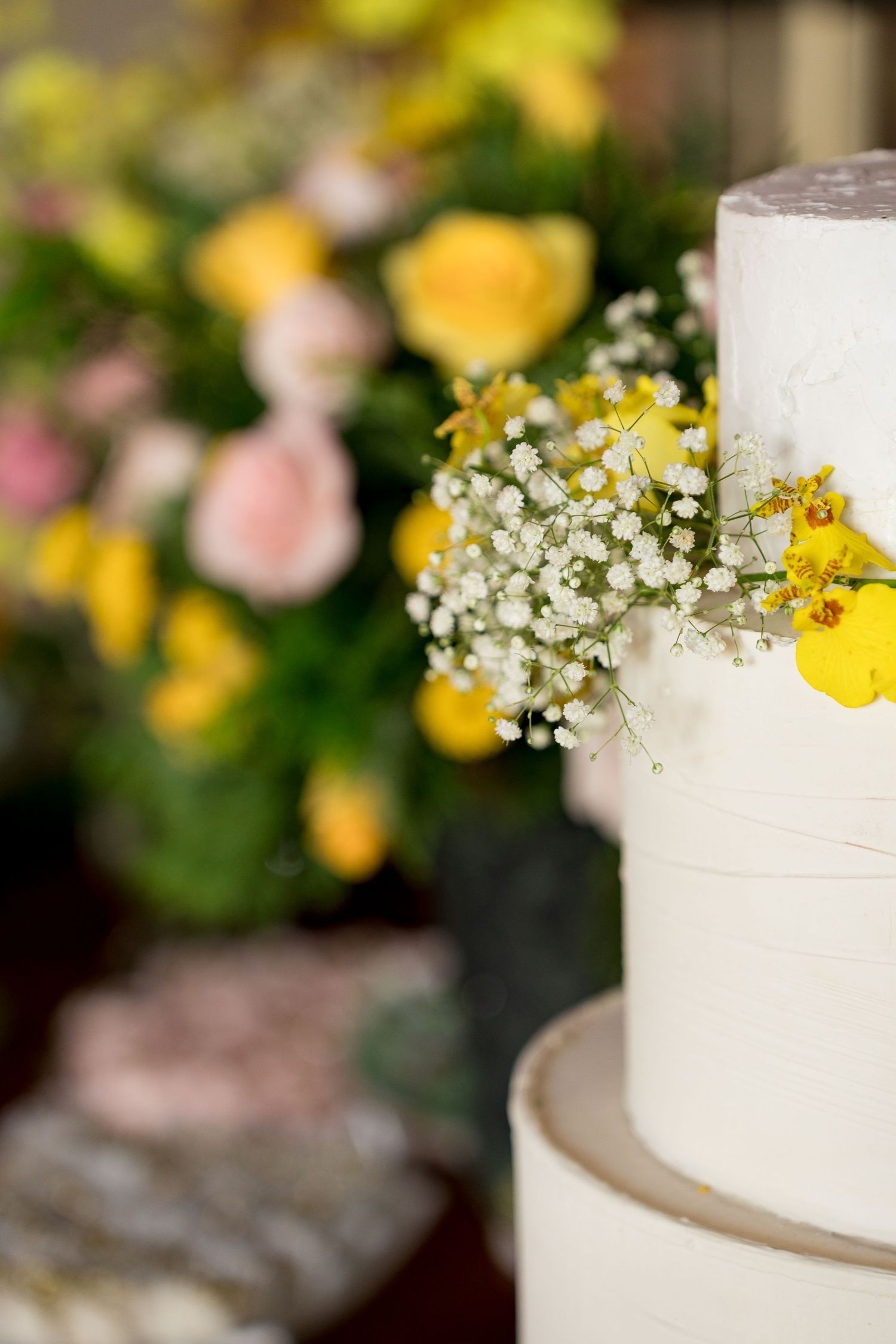 White tiered wedding cake adorned with yellow orchids and baby's breath, with colorful floral arrangement in background.
