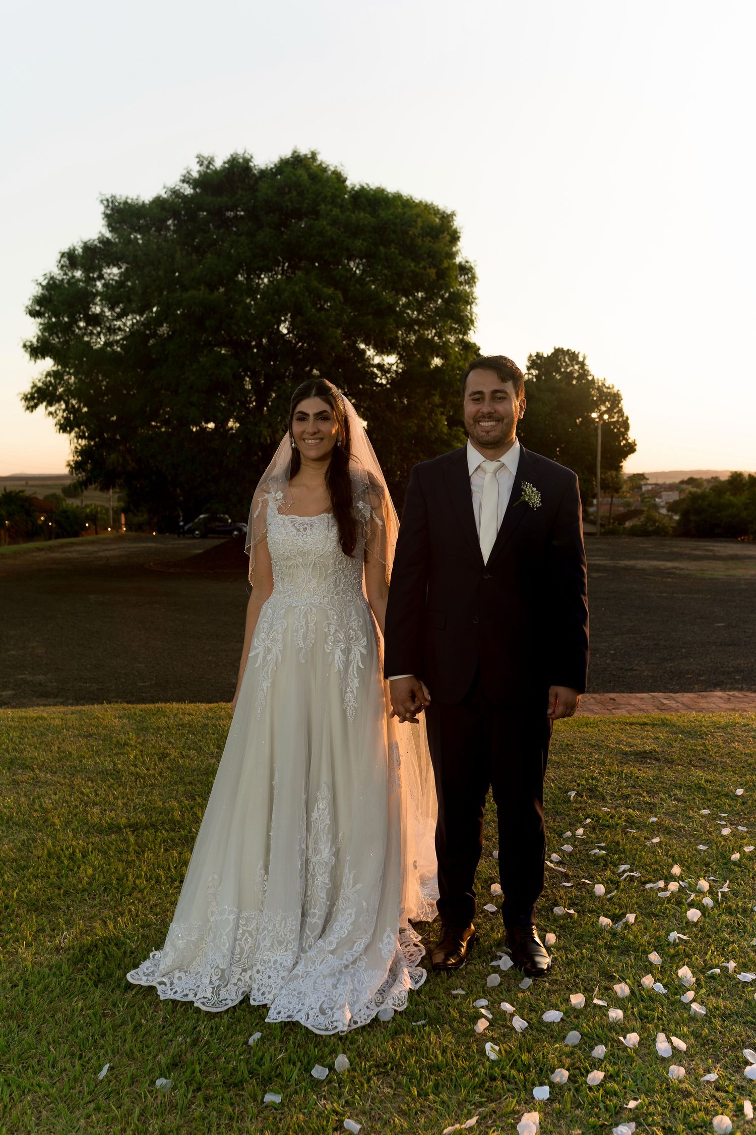 Bride in white lace gown and veil stands with groom in dark suit on green lawn at sunset near large tree.