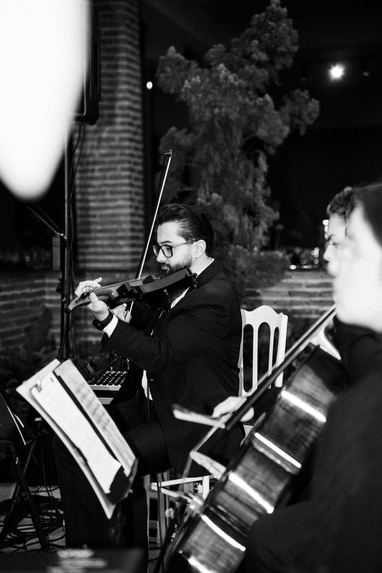 Black and white photo of a violinist in a suit performing at an outdoor evening concert with other musicians.