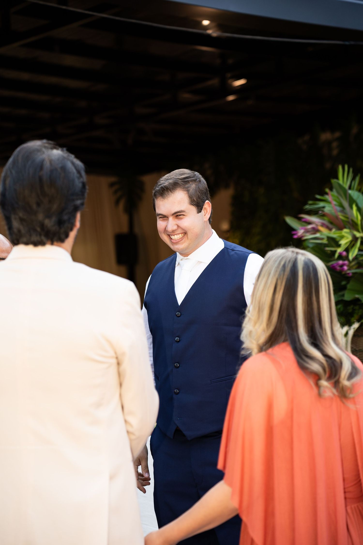 Smiling groom in navy blue vest and white shirt greeting wedding guests outdoors at elegant venue.