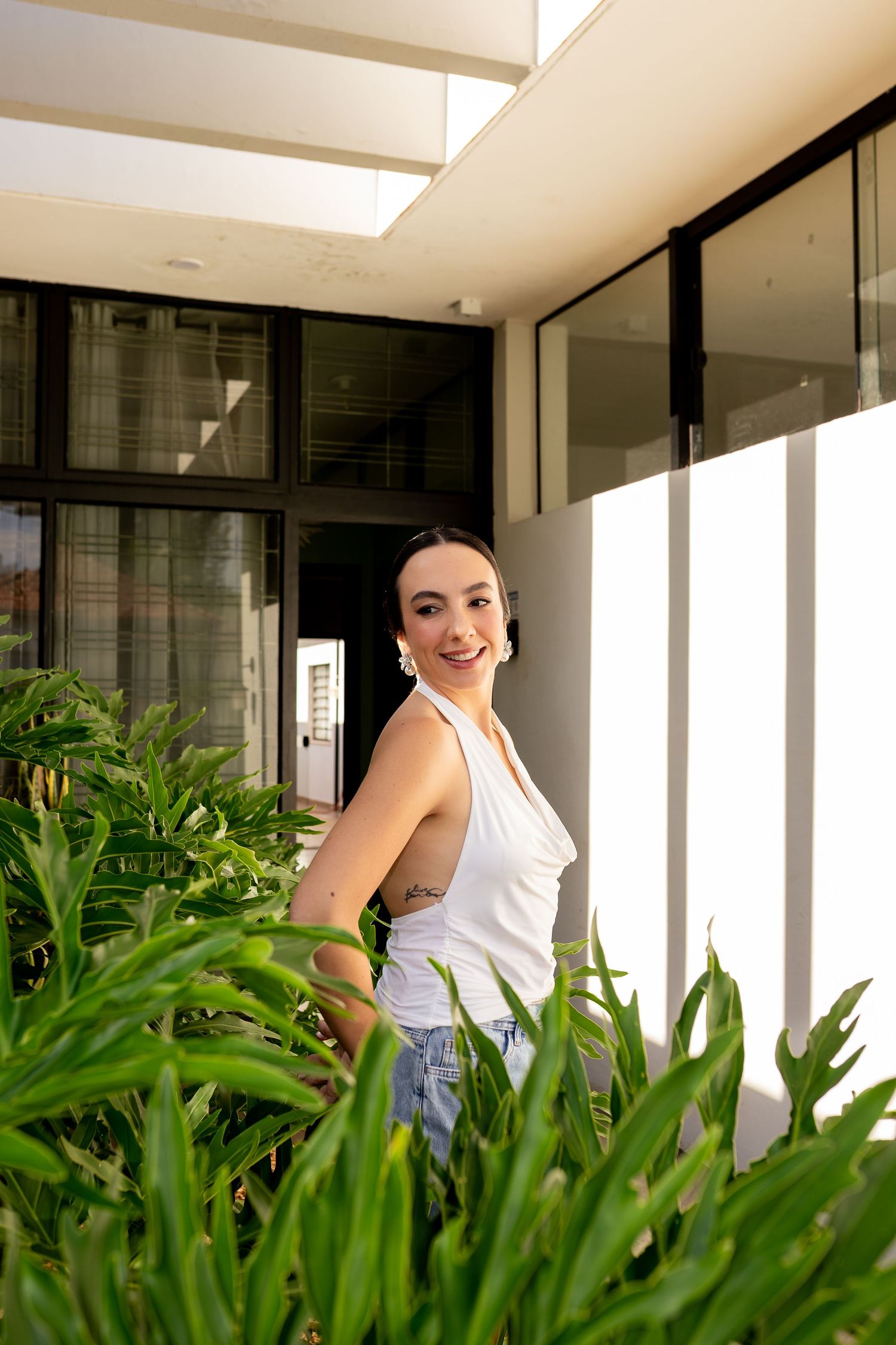 Young woman in white tank top and jeans smiles beside lush green plants outside a modern building.