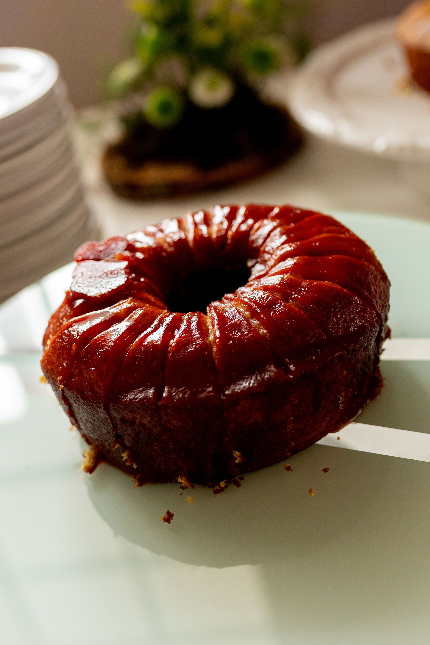 A glossy caramel bundt cake with rich brown glaze sits on a white plate, with stacked dishes in the background.