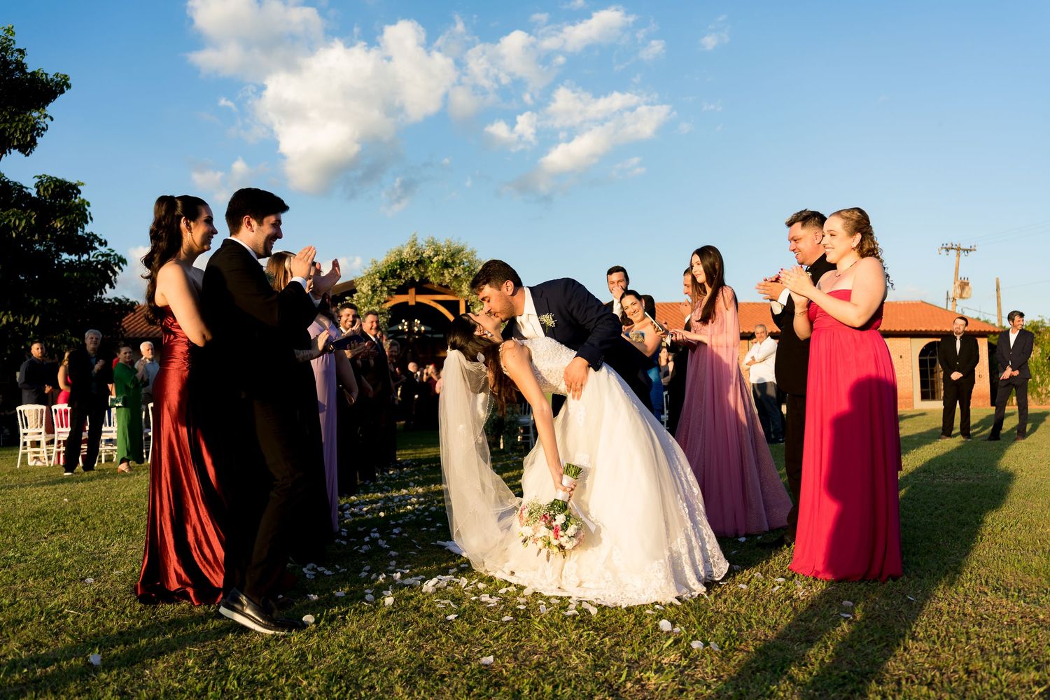Bride and groom share a kiss at outdoor wedding ceremony while bridesmaids in red and purple gowns applaud.