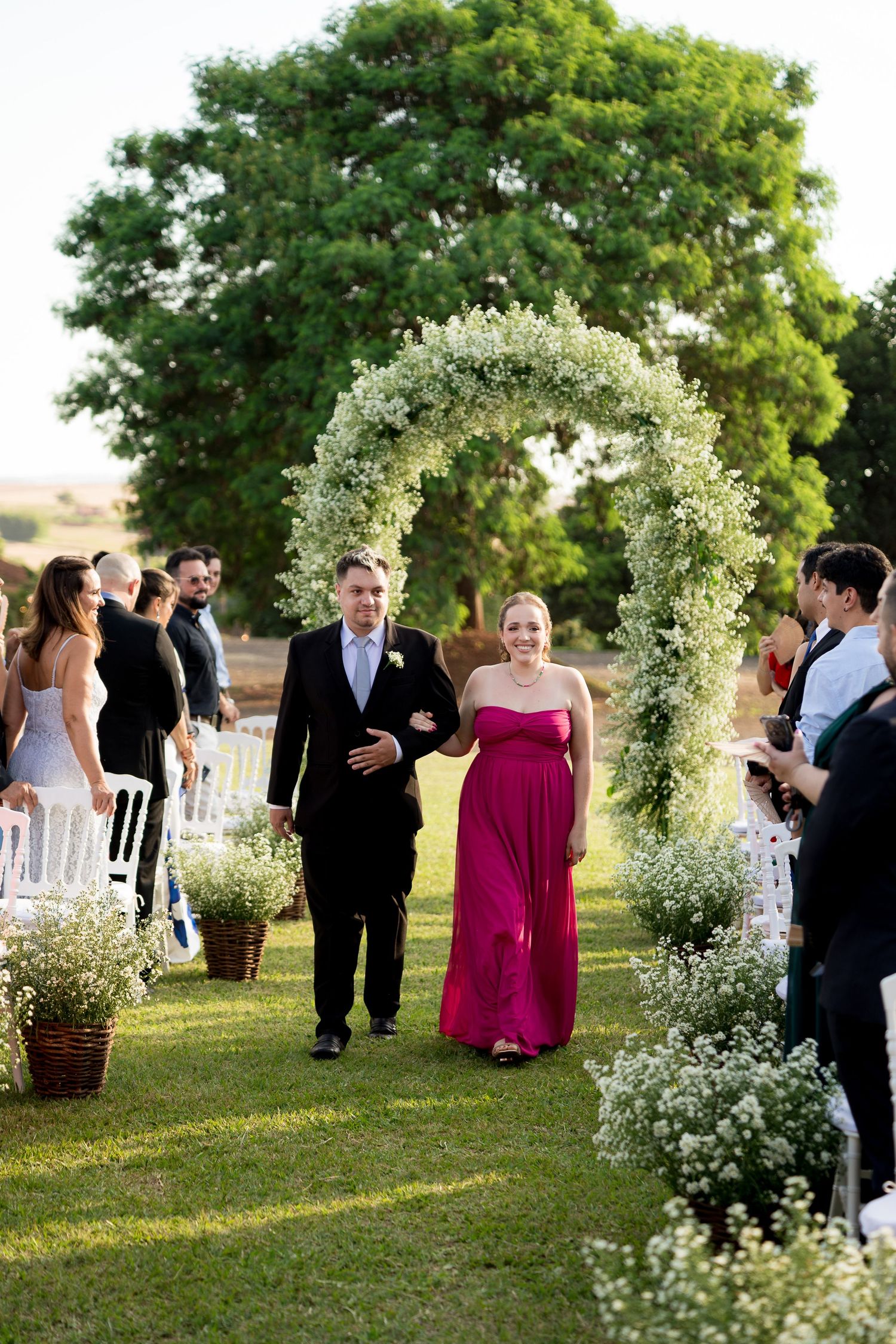 Couple walks down outdoor wedding aisle under floral arch, guests applaud, woman in pink gown, man in black suit.