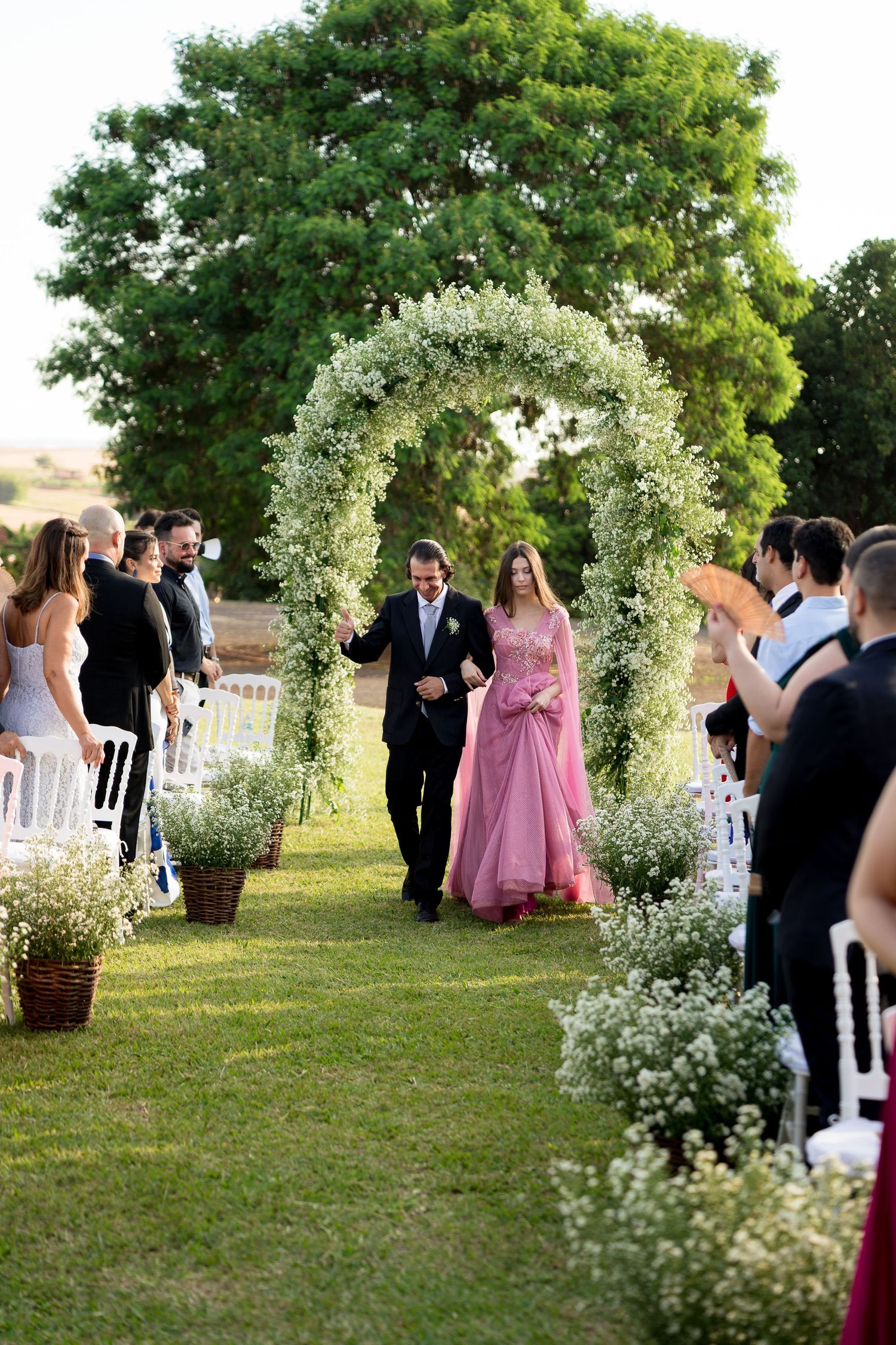 Couple walks down floral arch aisle at outdoor wedding ceremony, bride in pink gown, guests applauding on green lawn.