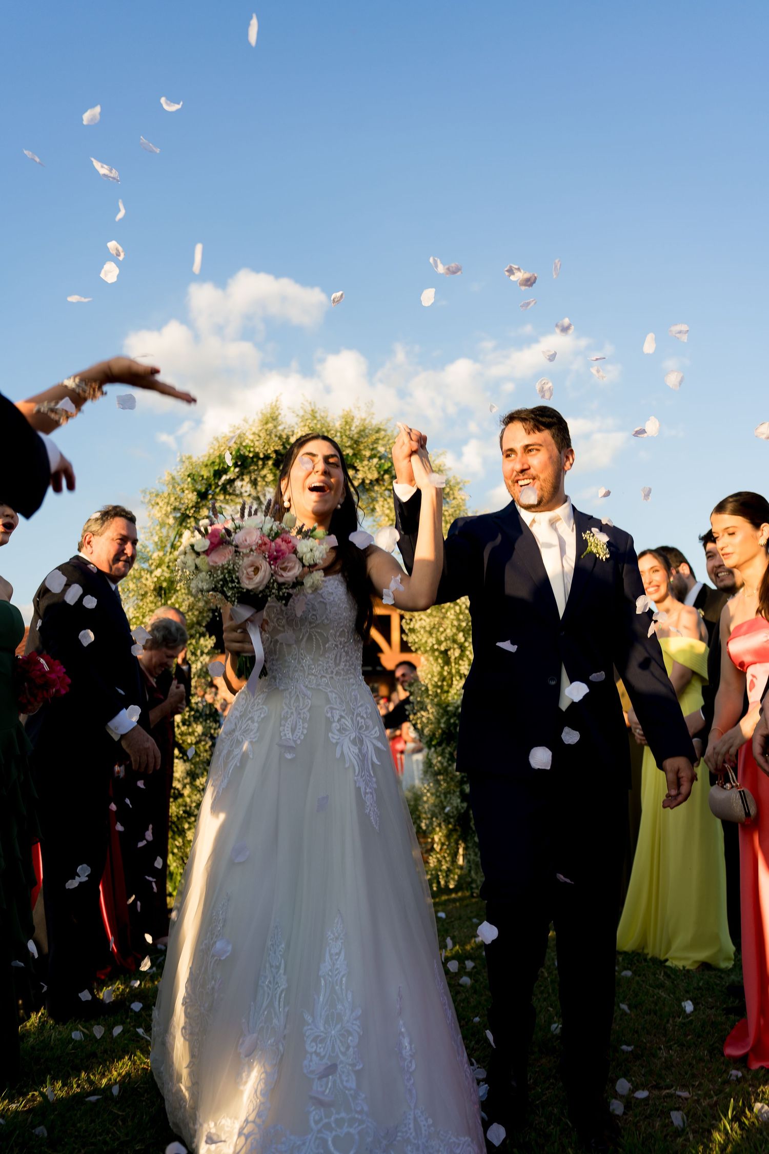 Joyful newlyweds walk through confetti shower, bride in lace gown holding bouquet, guests celebrating outdoors under blue sky.
