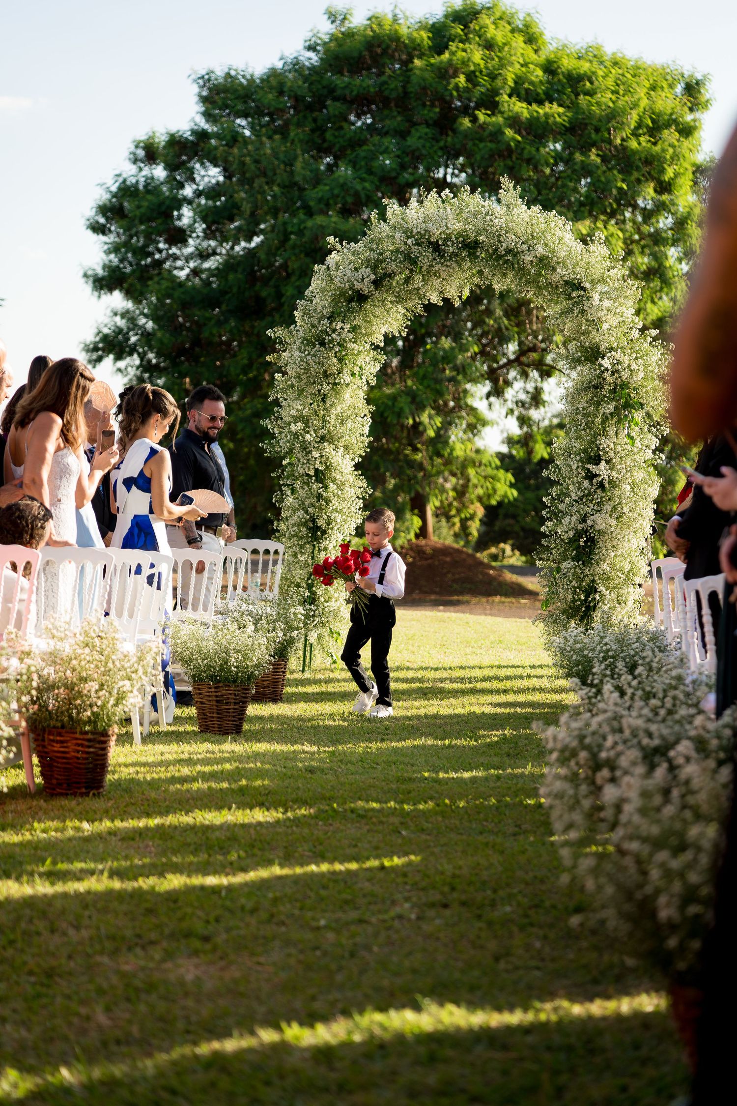 Child ring bearer walks down floral arch-adorned outdoor wedding aisle with red roses, guests seated on white chairs.