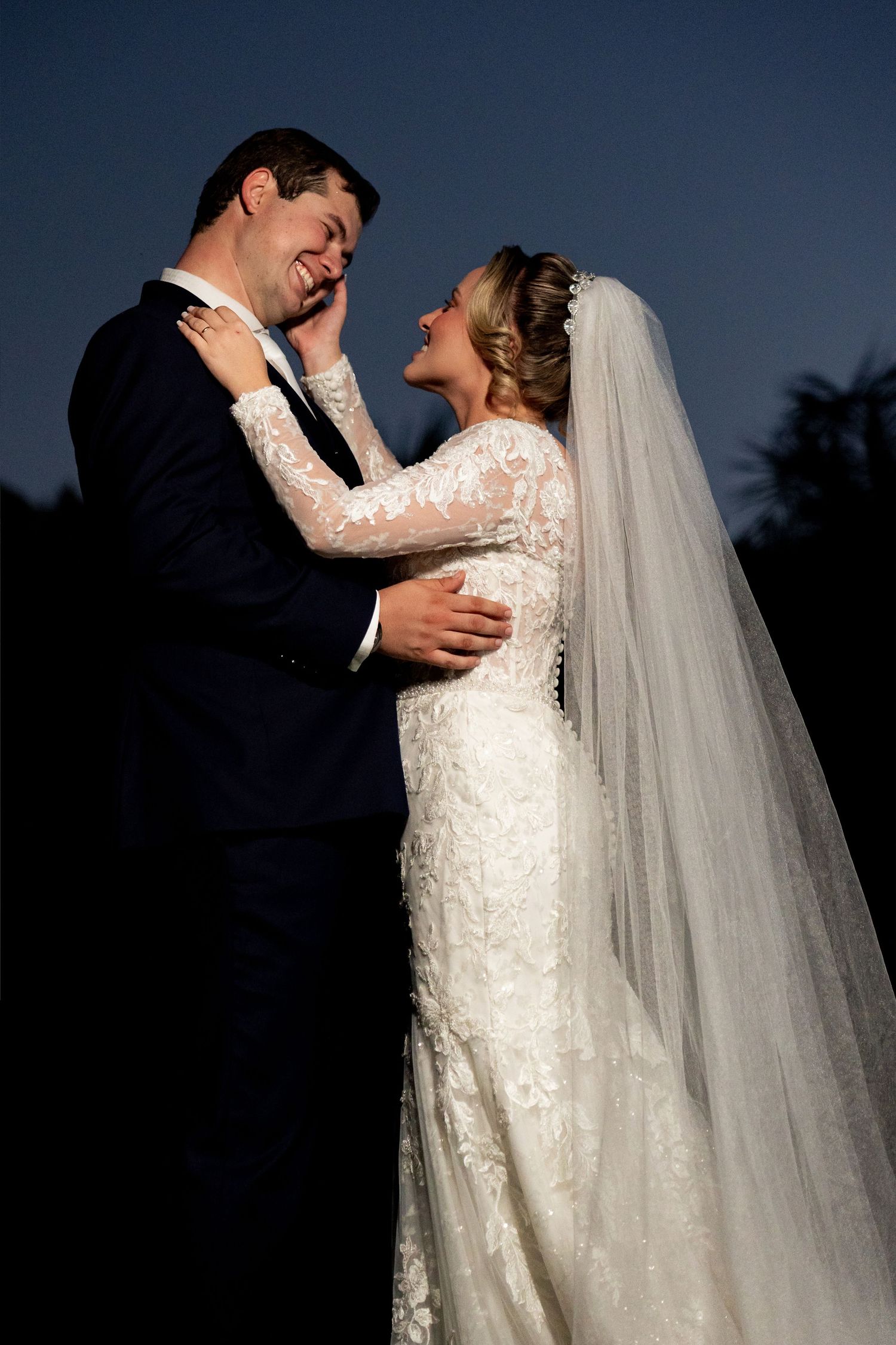 Bride in lace gown and veil laughs joyfully with groom in navy suit during romantic twilight wedding portrait.