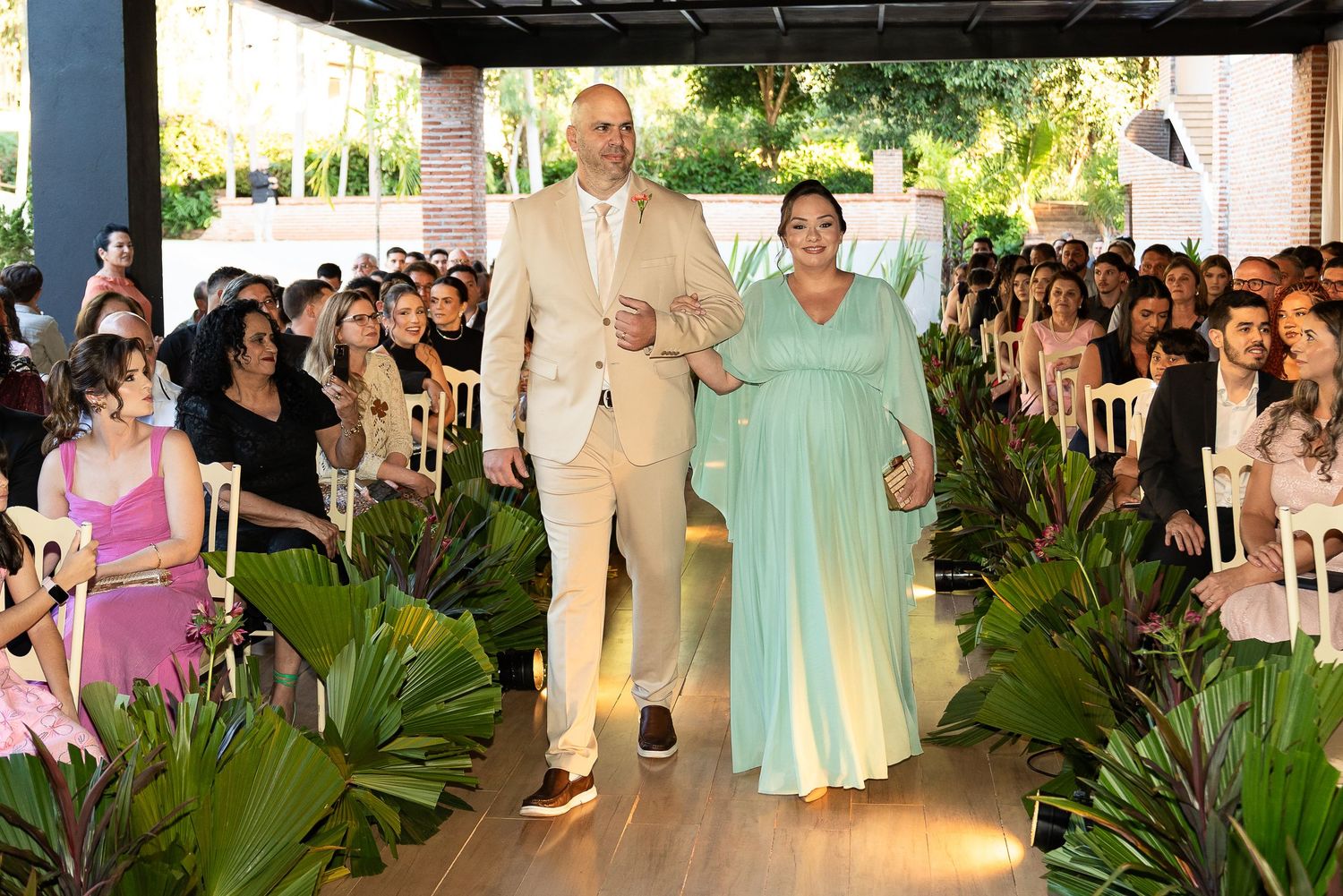 Couple walking down wedding aisle surrounded by tropical plants and seated guests at an outdoor ceremony.