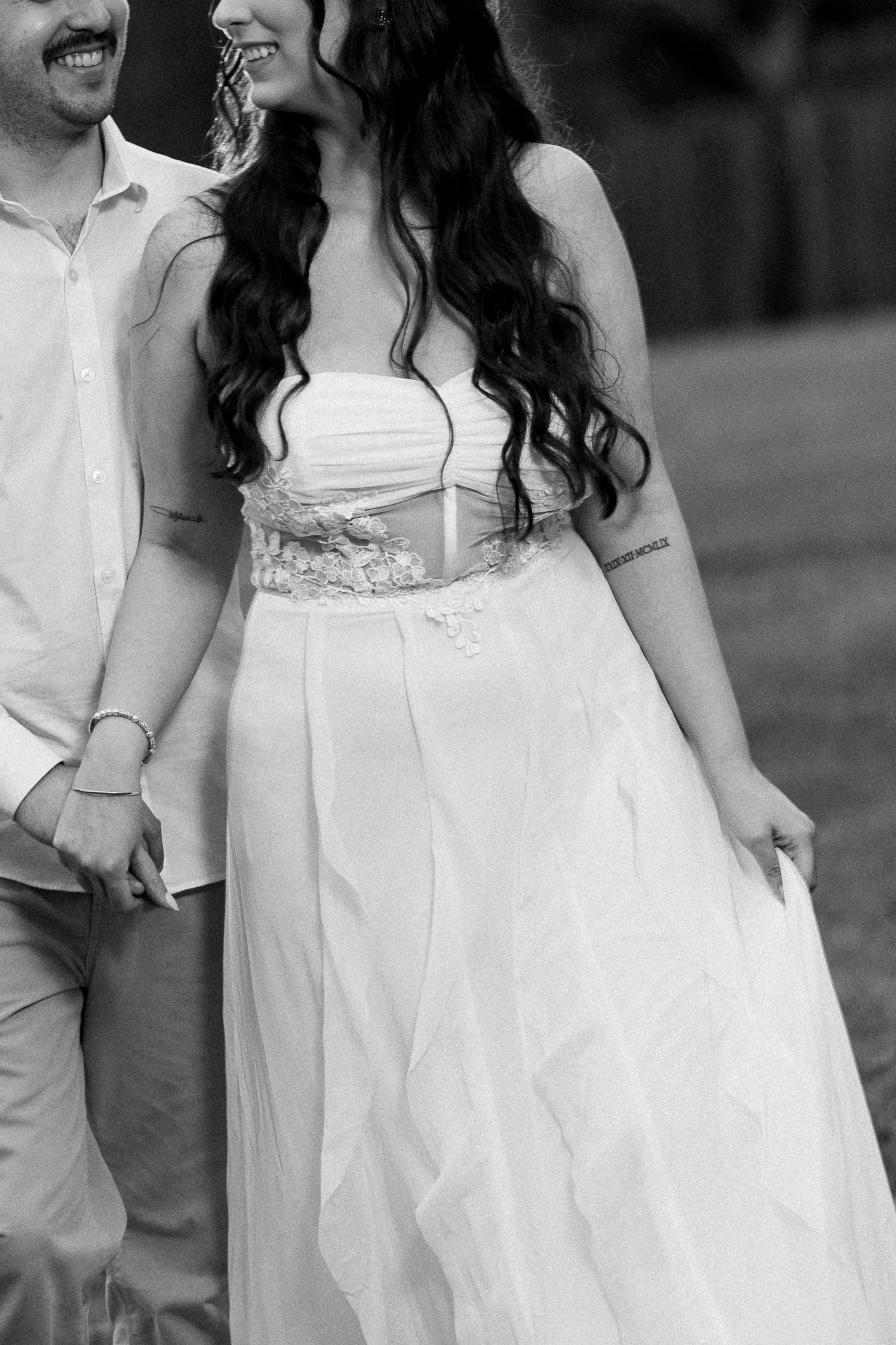 Black and white photo of a bride in a floral-belted wedding gown holding hands with her groom outdoors.