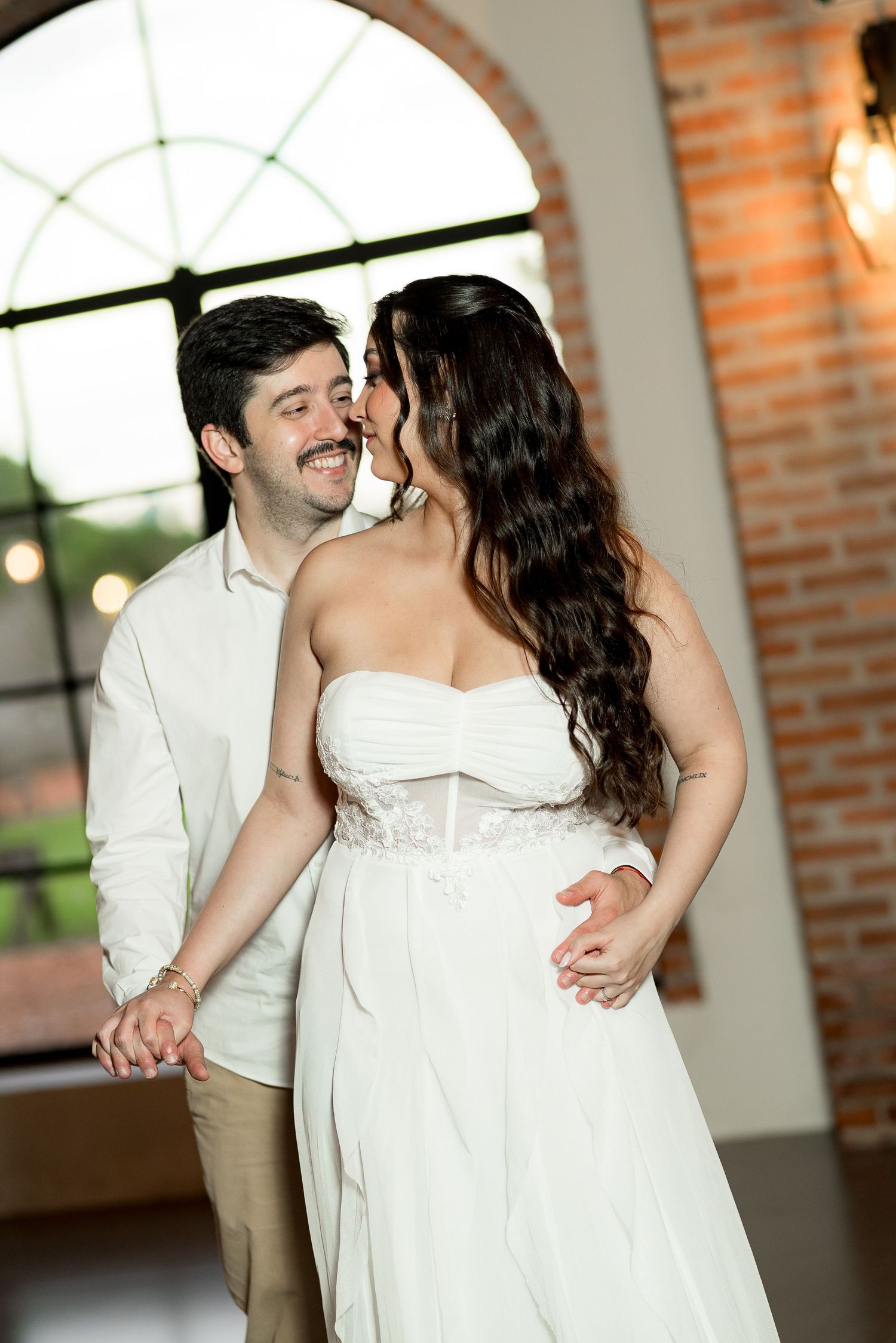 A smiling groom in white shirt holds his bride in a strapless white wedding gown as they share a tender moment indoors.