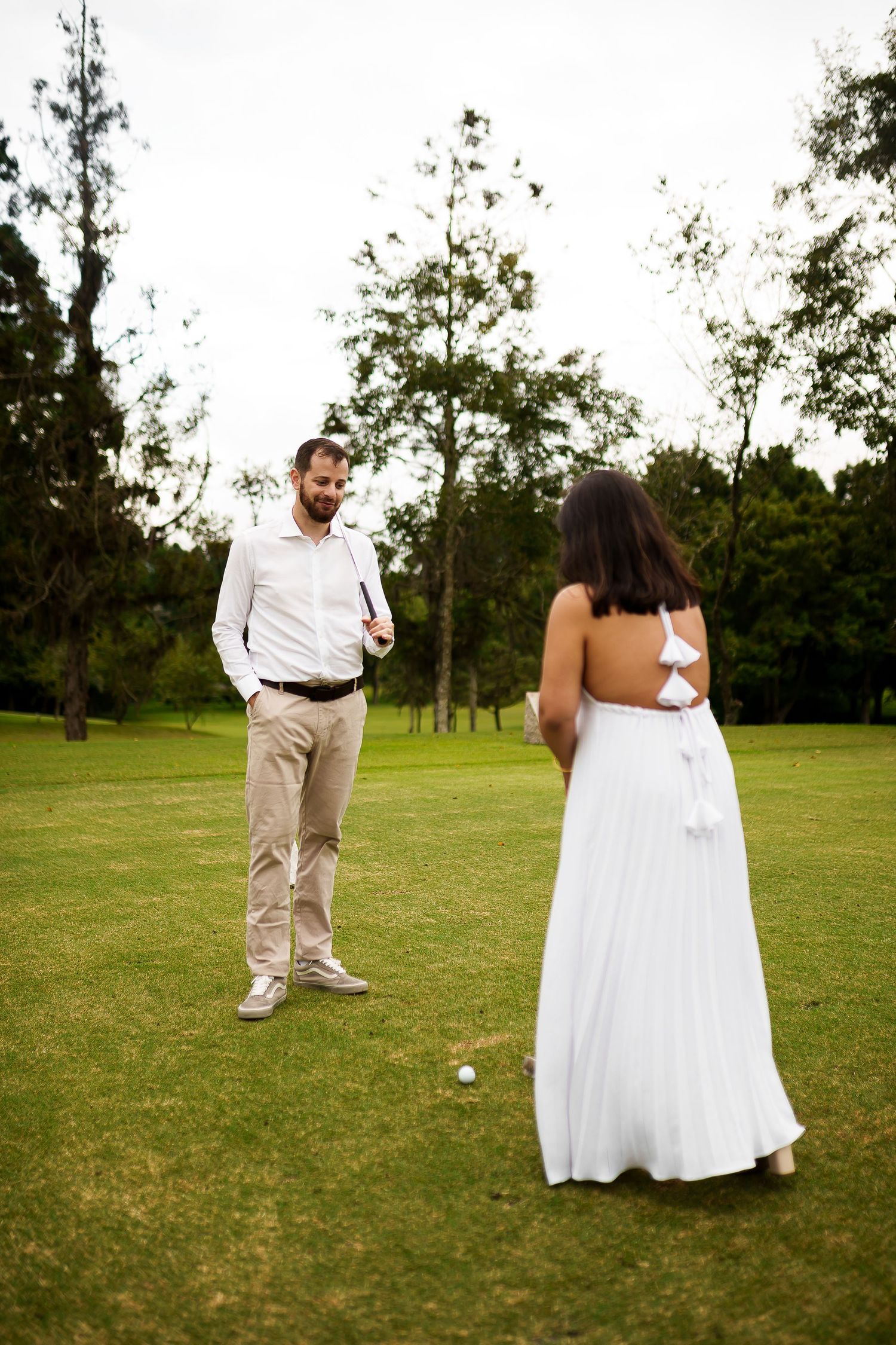 Couple in white attire playing golf on a lush green course, woman in backless dress, man holding a club.
