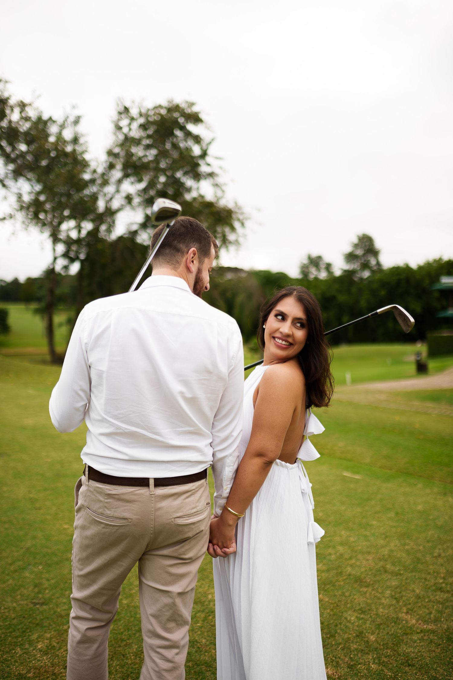 Couple holding hands on a golf course, woman in white dress smiling back at camera, man in white shirt holding a golf club.