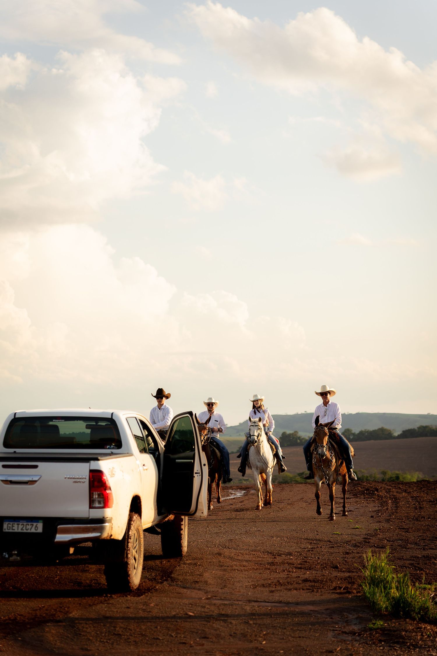 Three cowboys on horseback ride down a dirt road past a white pickup truck under a cloudy sky.