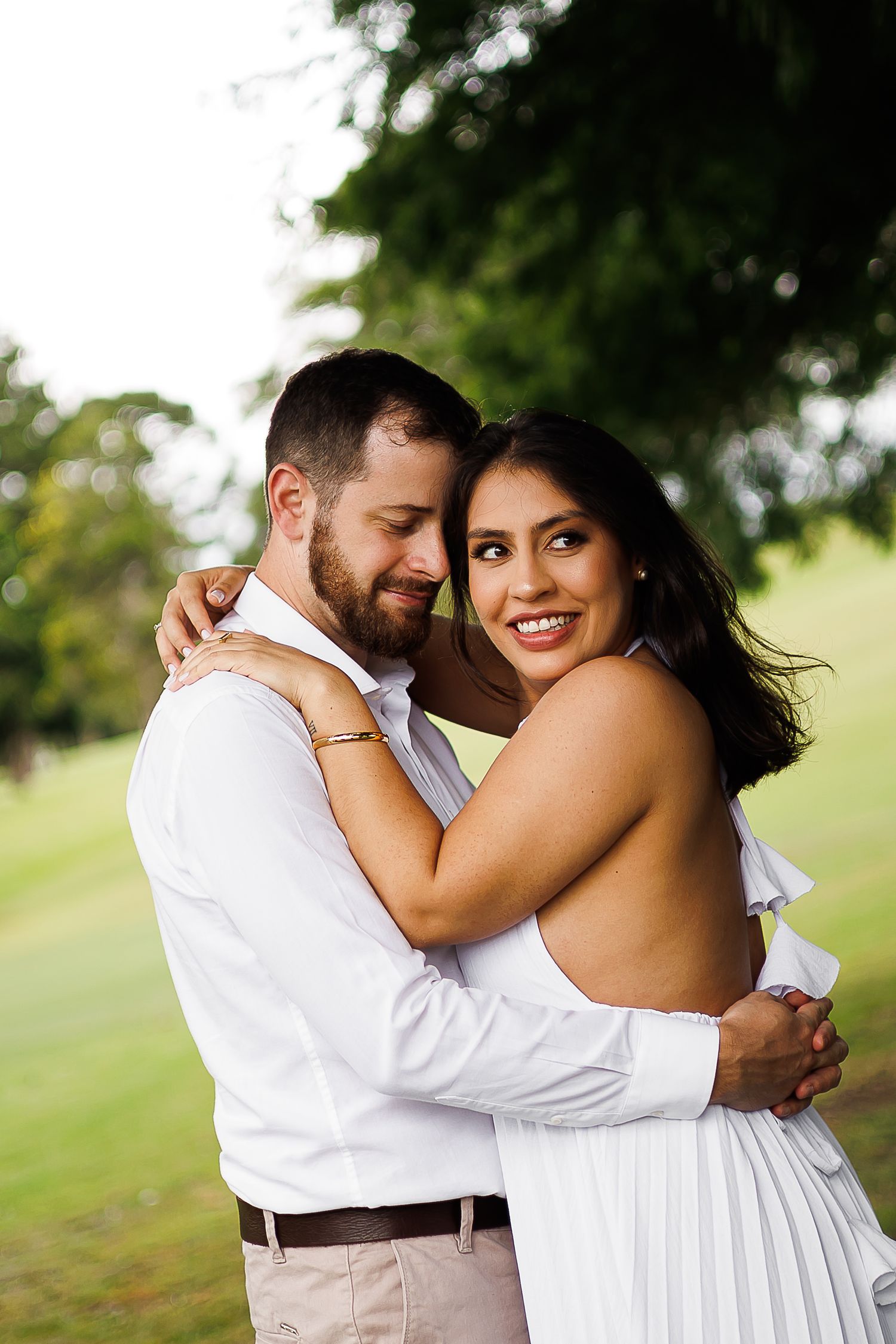 Couple embracing outdoors in white outfits, smiling warmly, with lush green trees in the background.