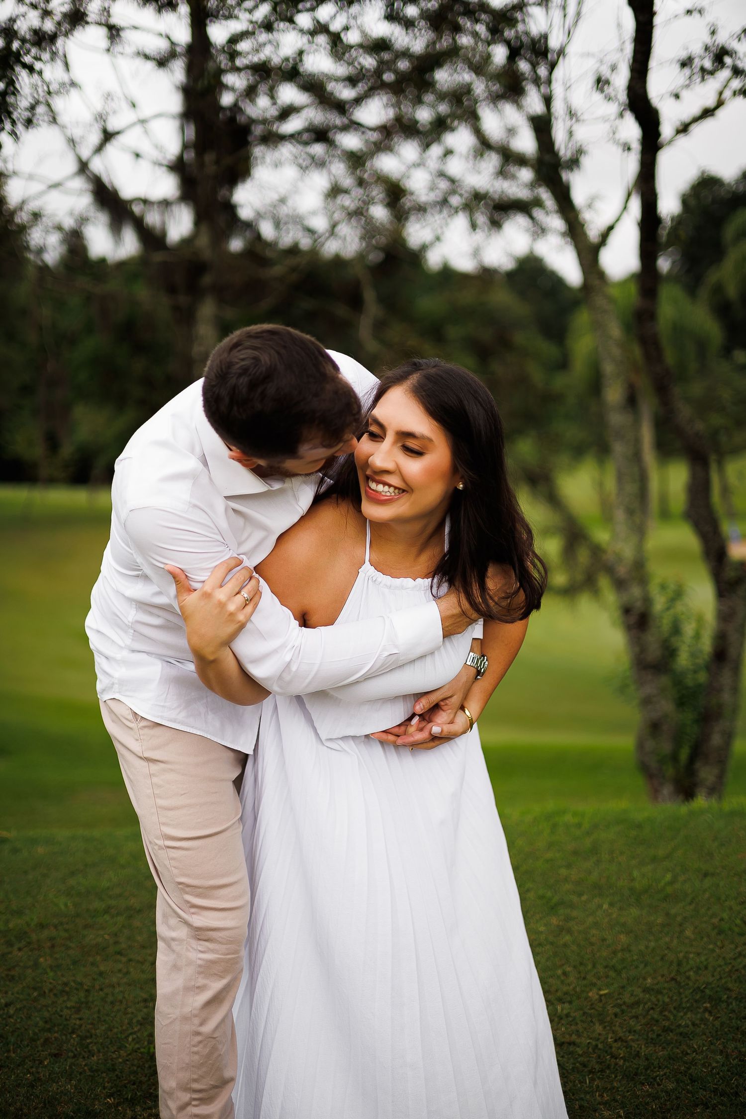 A couple in white outfits embracing on a green lawn, the woman smiling joyfully as the man nuzzles her neck.