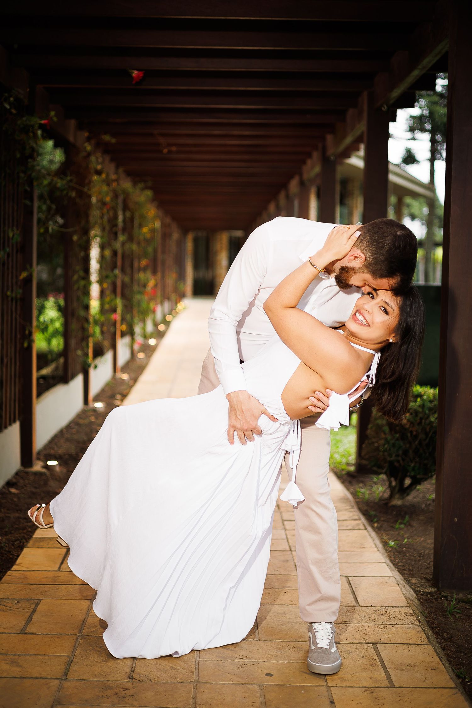 Groom dipping bride in white dress under wooden pergola, sharing romantic kiss at outdoor wedding venue.