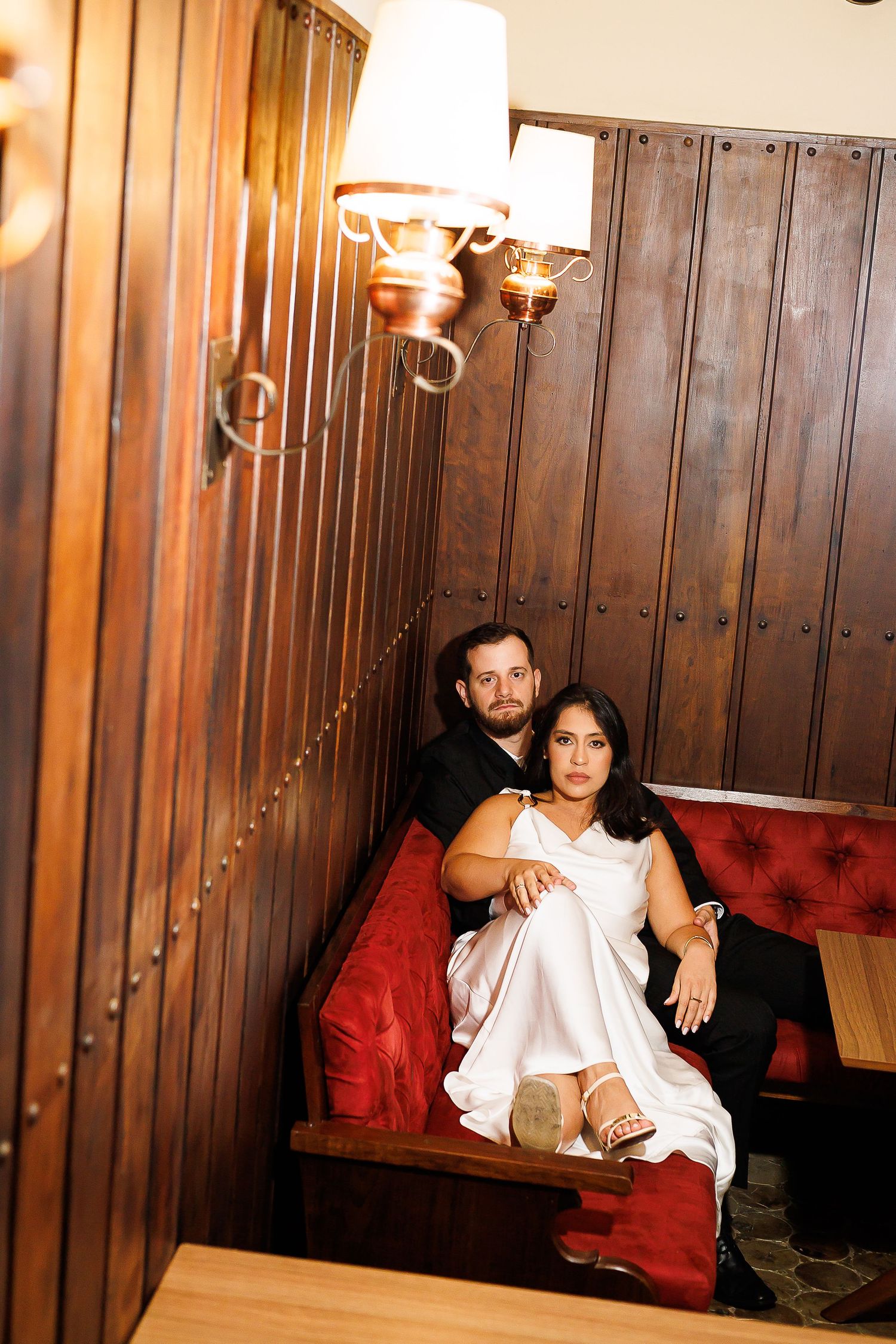 Couple posing in a cozy restaurant booth with red velvet seats and warm wood paneling, woman in white dress.