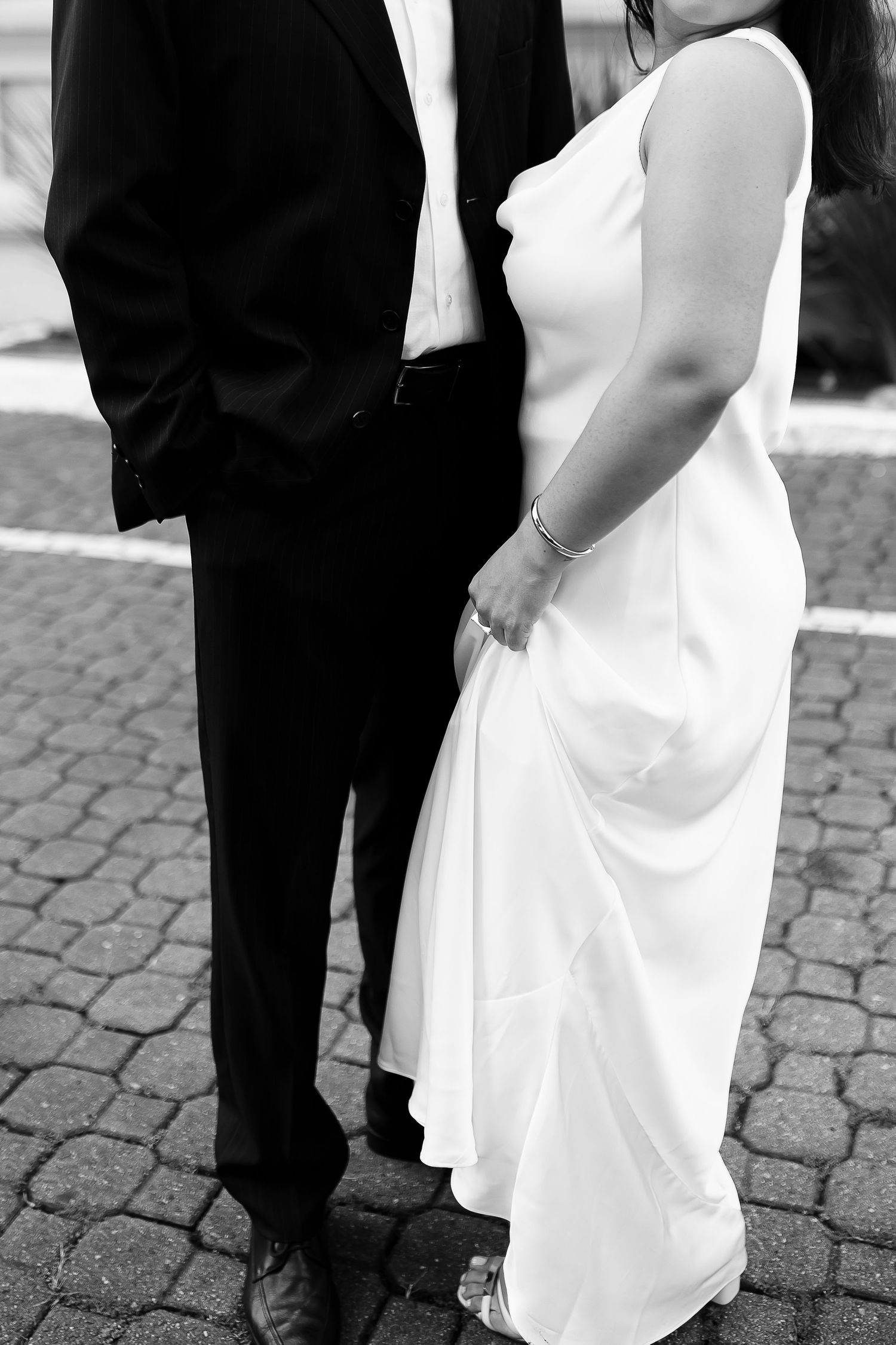 Black and white photo of a bride in white gown and groom in tuxedo standing on cobblestone street.