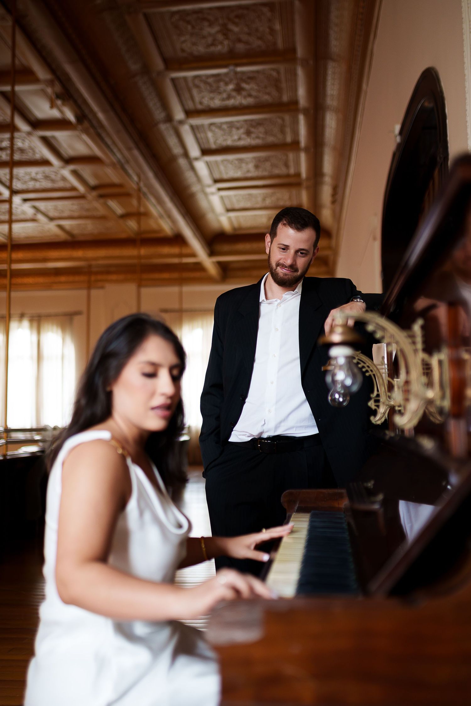A woman in a white dress plays piano while a man in a black suit leans on it, holding a glass in an elegant room.